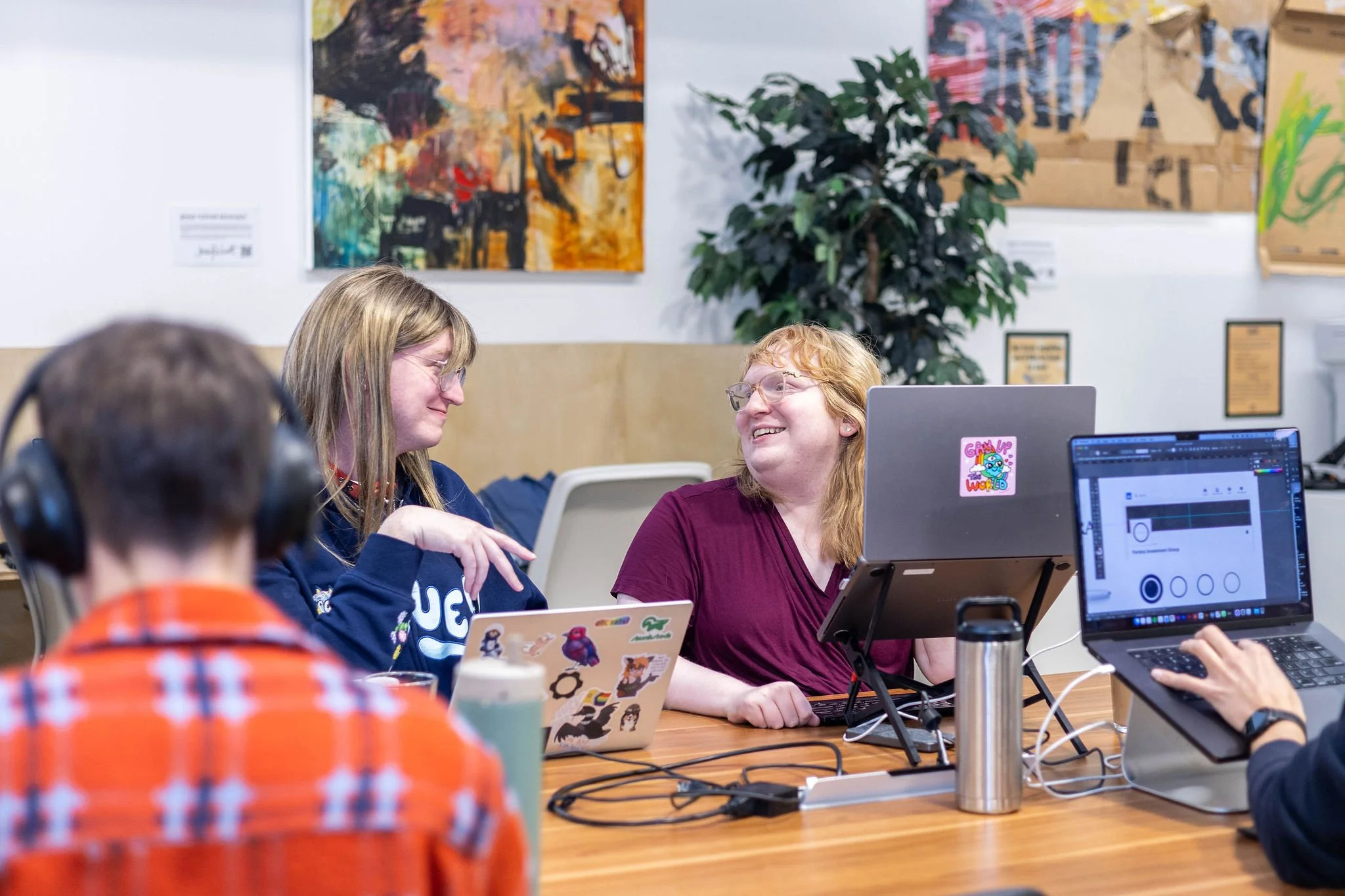 Four people in a room with artwork on the walls and a large plant. Two women are smiling and talking to each other. A person in the foreground with headphones is working on a laptop, and another person on the right is using a laptop with a design pro