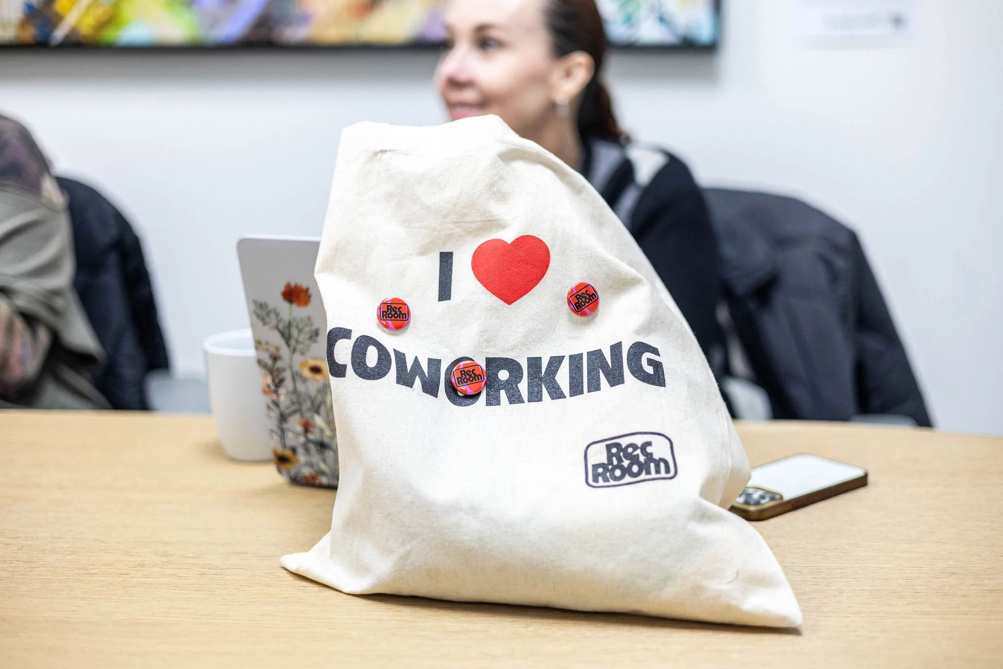 A beige tote bag with the text 'I ❤️ COWORKING' and the REC ROOM logo, placed on a table in a meeting room. Two women are in the background, one with a laptop and the other with a backpack.