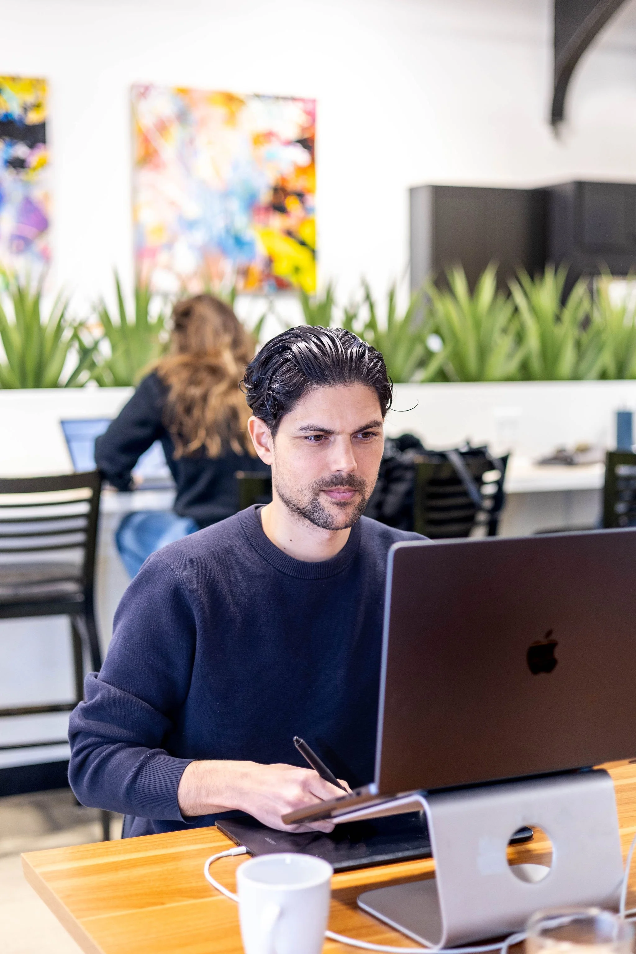 A man with dark hair and a beard working on a laptop at a wooden table in an office or coworking space, with green plants and colorful abstract artwork on the white walls behind him.