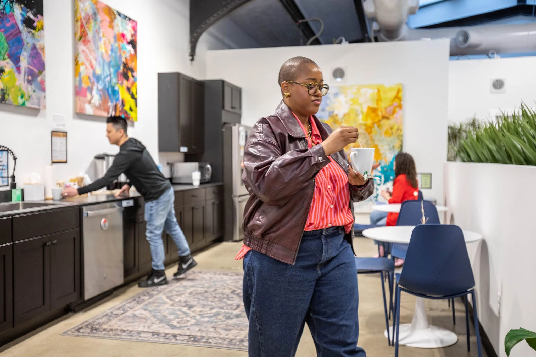 Woman with a bald head, glasses, and gold earrings, wearing a brown leather jacket and red striped shirt, holding a cup of coffee in a cafe.