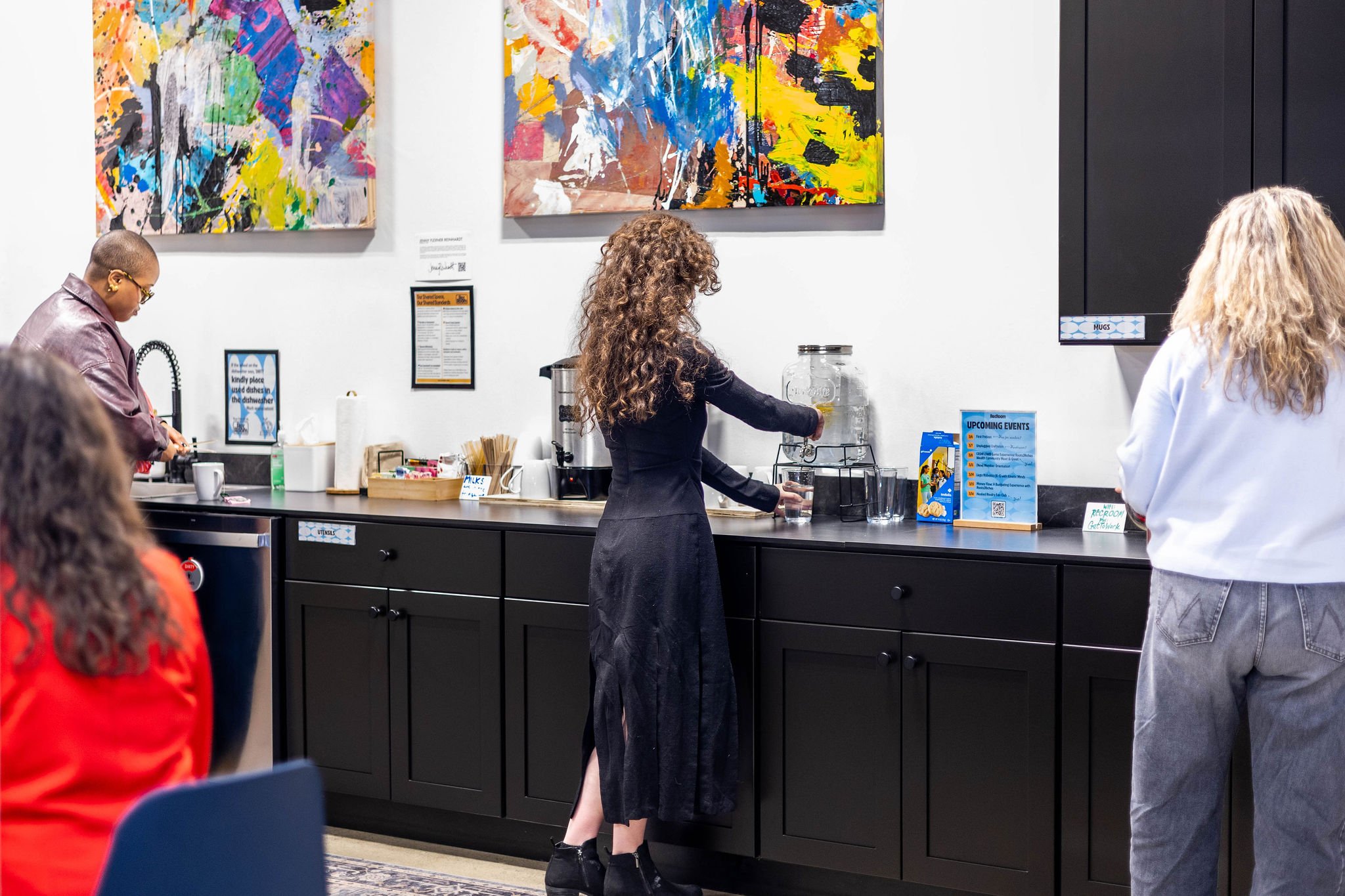 People gather around a black countertop in a gallery, with colorful abstract paintings on the white wall behind. A woman with curly hair and black attire fills a glass from a drink dispenser, while others wait or prepare food. There are informational