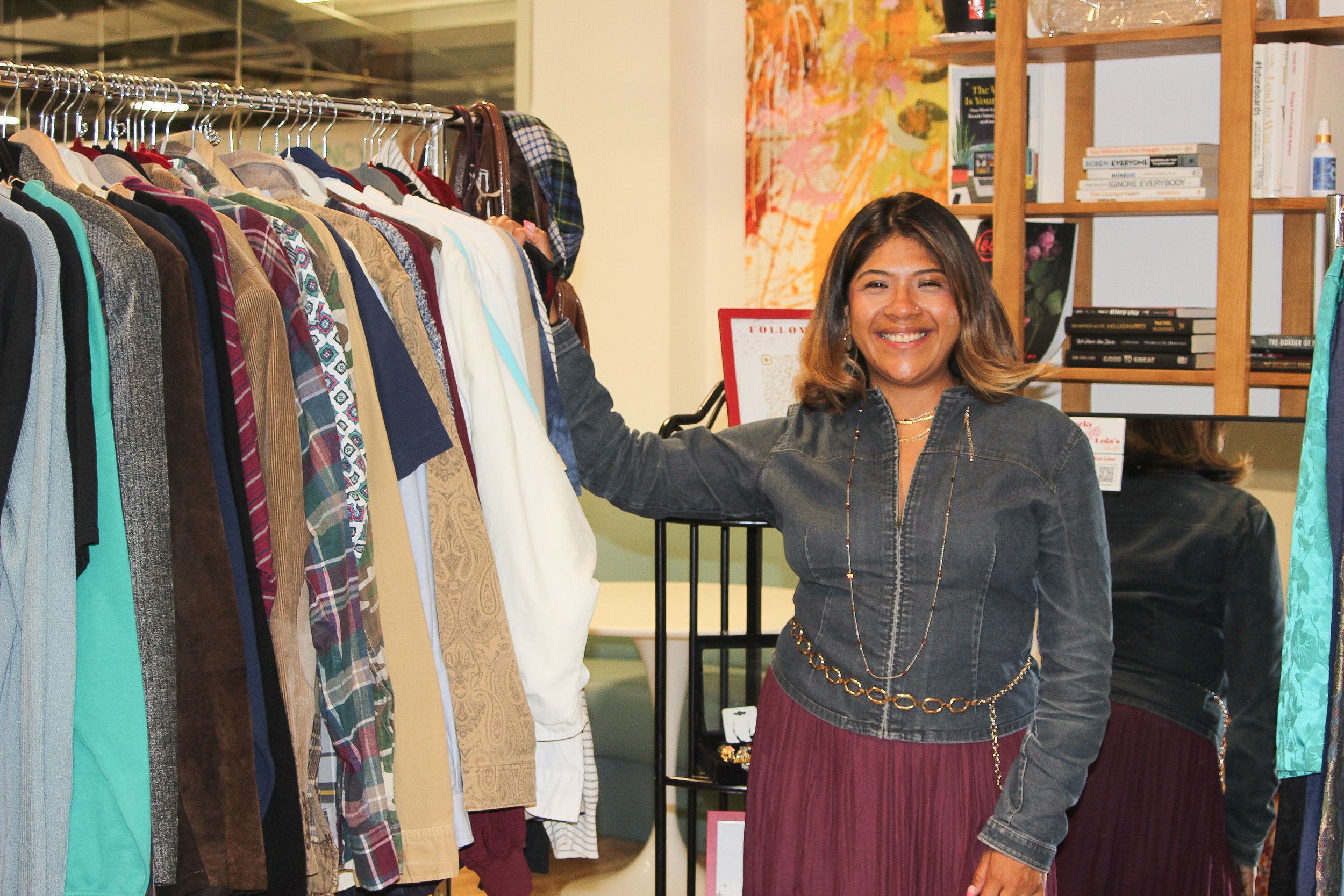 A woman shopping for clothes in a store, smiling, standing beside a rack of colorful men's shirts. She has shoulder-length hair, a nose piercing, and is wearing a denim jacket, a long necklace, and a burgundy skirt.
