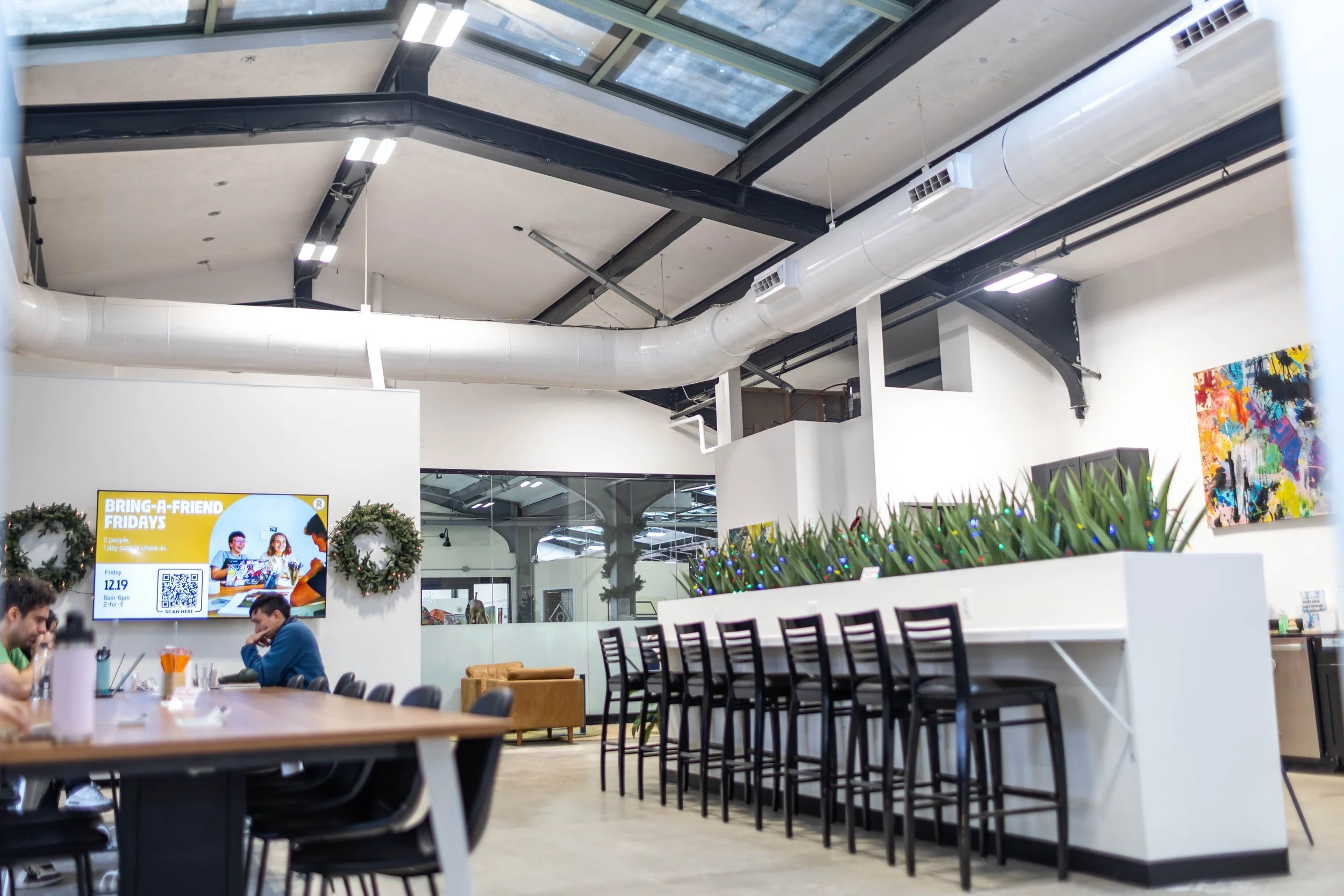 Modern indoor space with high ceiling, skylight, white walls, bar-height chairs, and artwork, decorated with holiday wreaths, and a large digital screen displaying an advertisement.