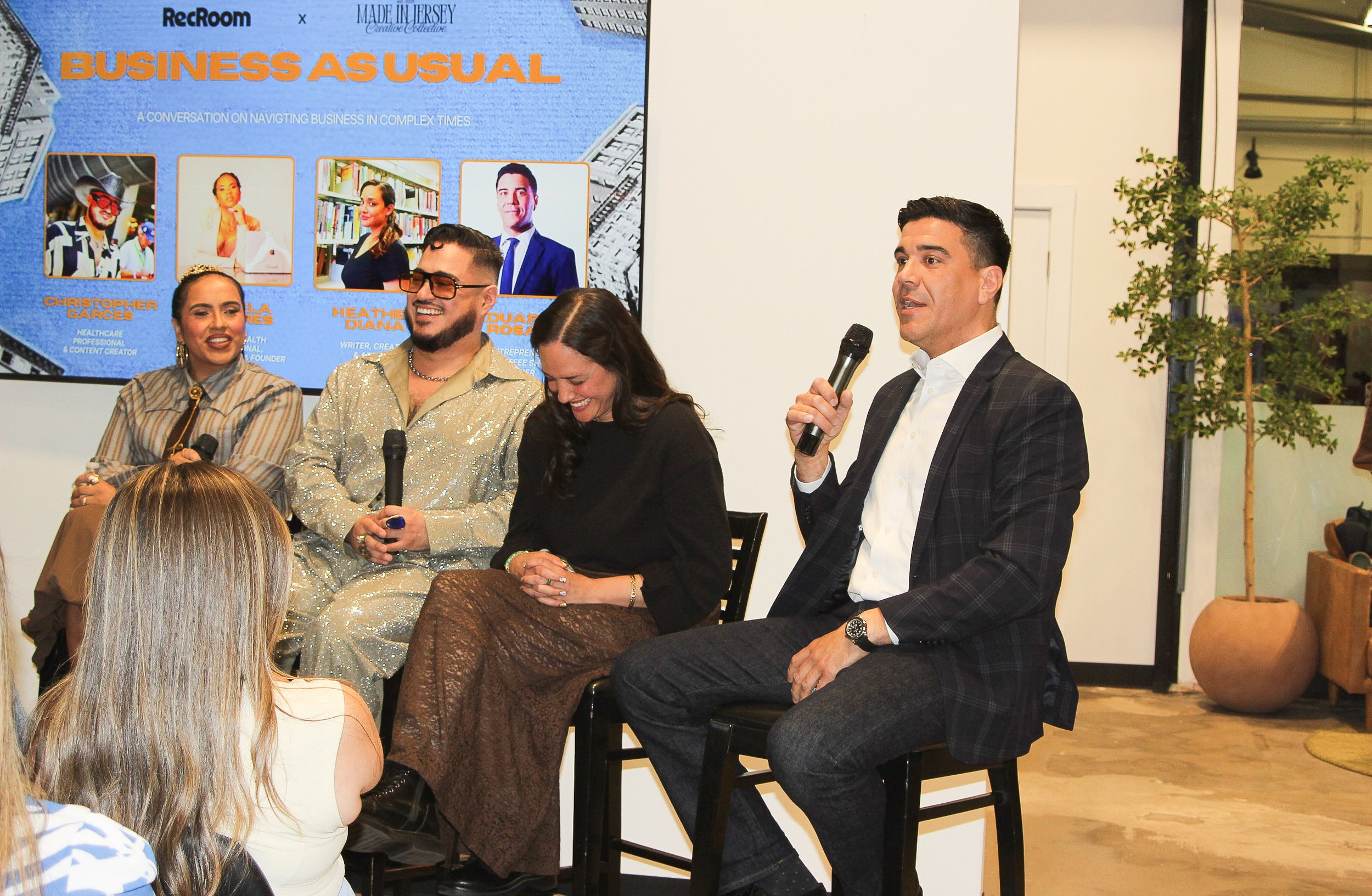 A man in a white shirt and checkered blazer speaking into a microphone during a panel discussion, with three women also seated and smiling, and a large screen behind displaying a presentation titled 'Business As Usual' with photos and names of four speakers.