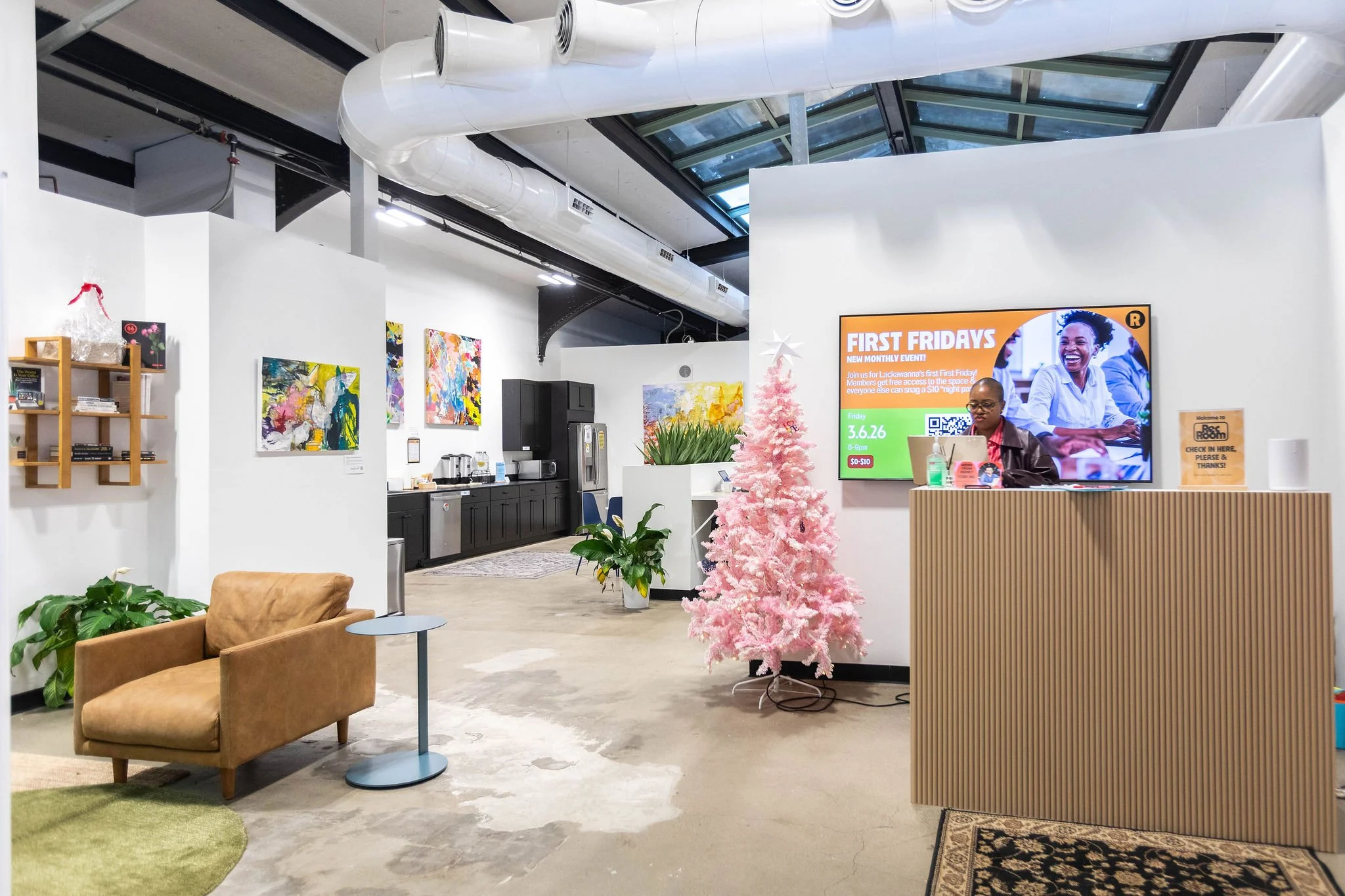 Interior of an office with a white Christmas tree, a reception desk, artwork on the walls, a seating area with a tan chair, plants, and a large digital screen displaying a promotional event.