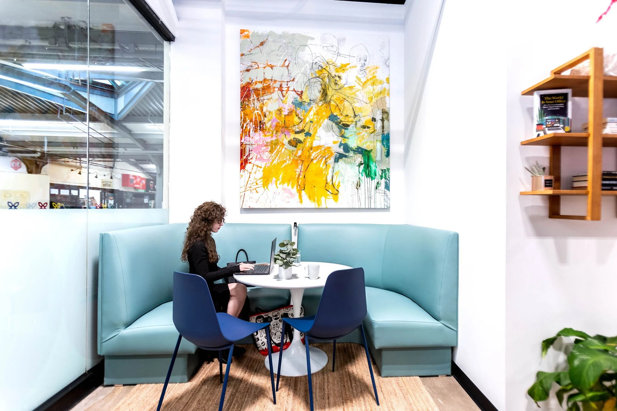 A woman with curly hair sitting at a round white table working on a laptop in a modern cafe or office space with teal booth seating, a colorful abstract painting on the white wall, and a wooden shelf with books.