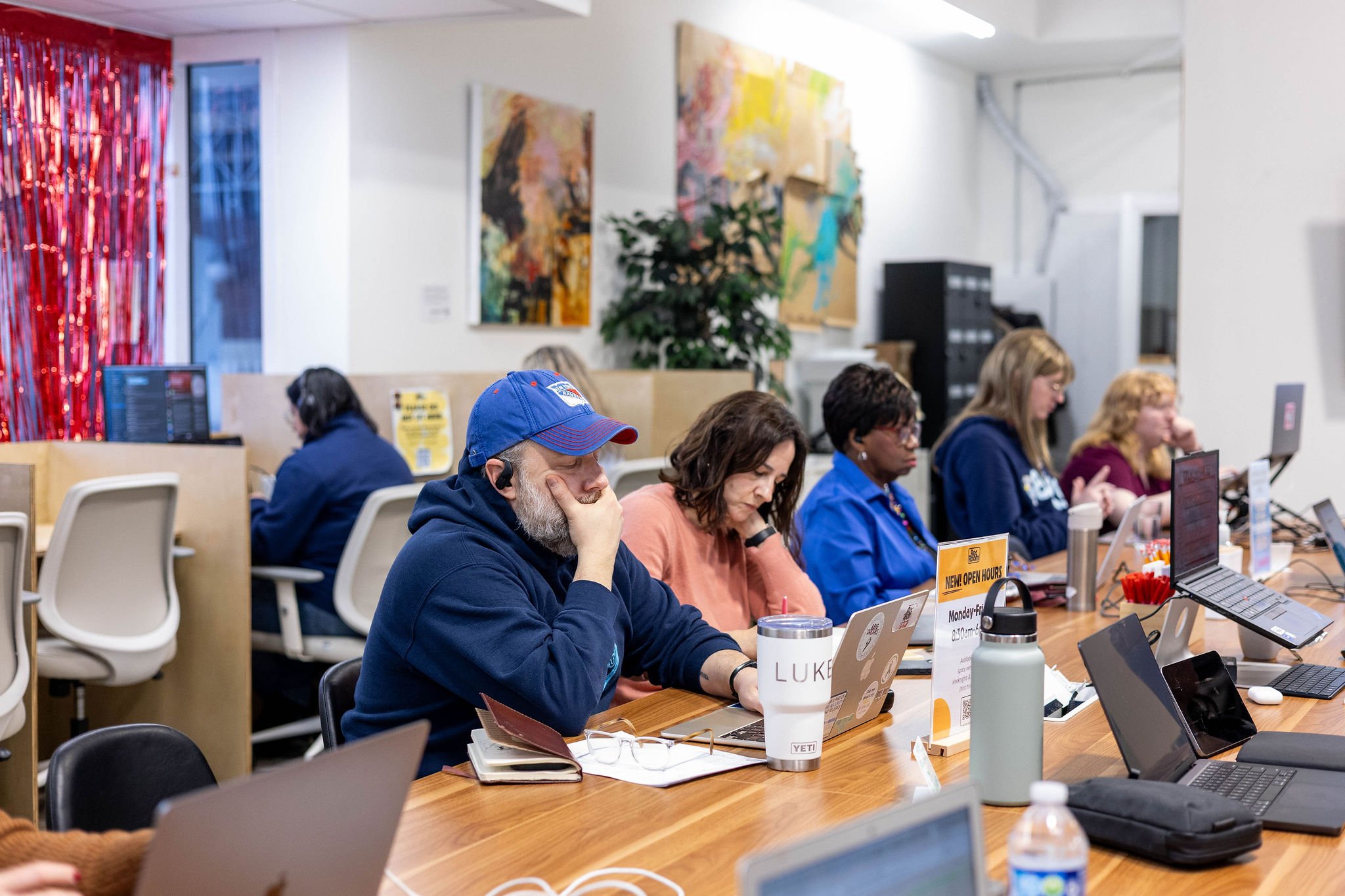 People working on laptops at a long table in a modern office or co-working space, some of whom are focused and some using headphones, with colorful artwork on the walls and desks with various beverages and supplies.