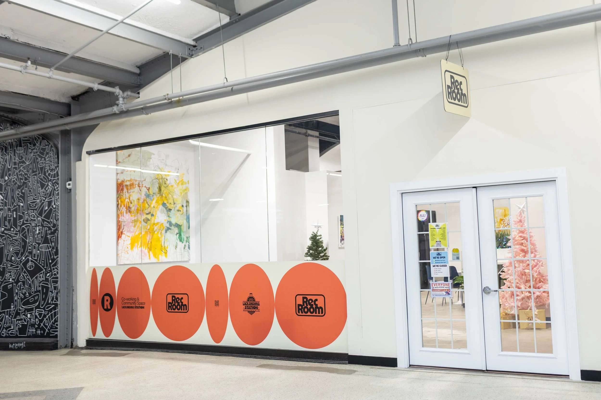 Entrance to a community space or coworking space with a large glass window, white walls, and pink Christmas tree inside, decorated with colorful art on the wall and signs on the door.