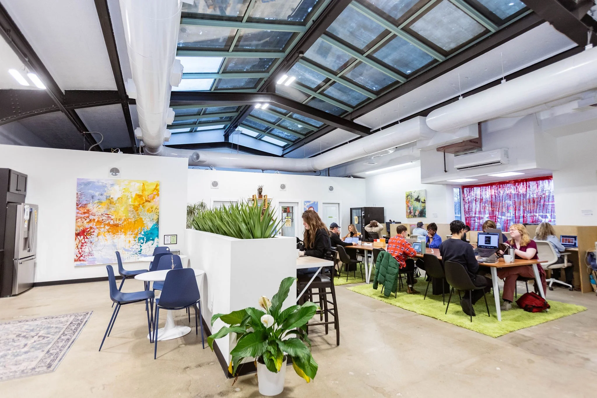 Interior of a modern coworking space with people working on laptops at tables, colorful artwork on the walls, plants, and large windows with a glass ceiling.