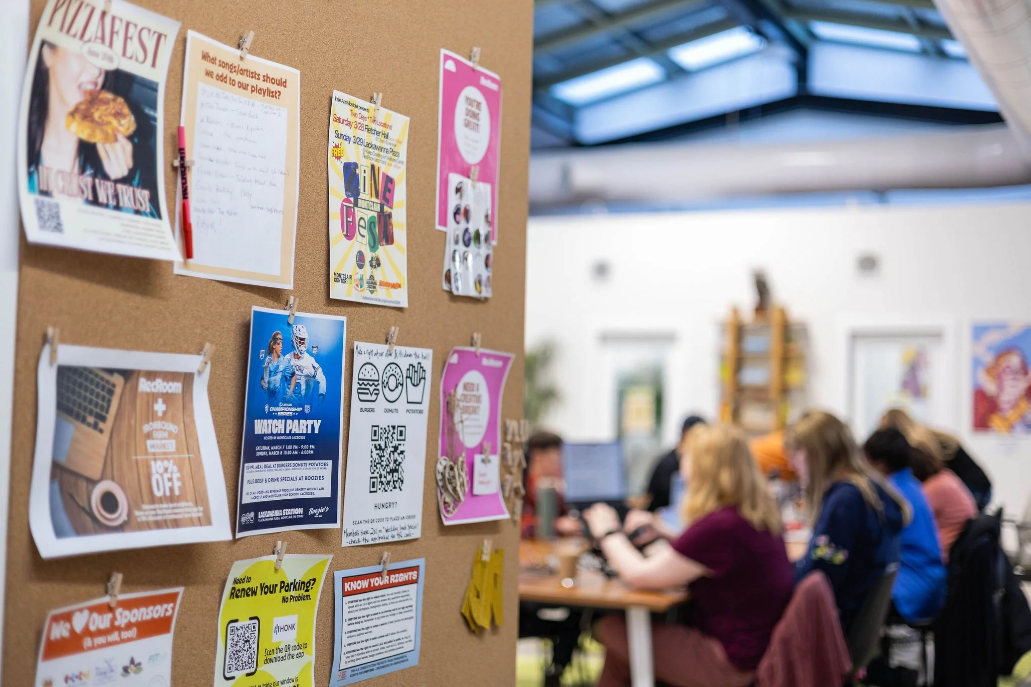 A cork bulletin board with various flyers and notices, including an event poster and advertisements. In the background, a group of people sit at a long table, working on laptops in a well-lit indoor space with a glass ceiling.