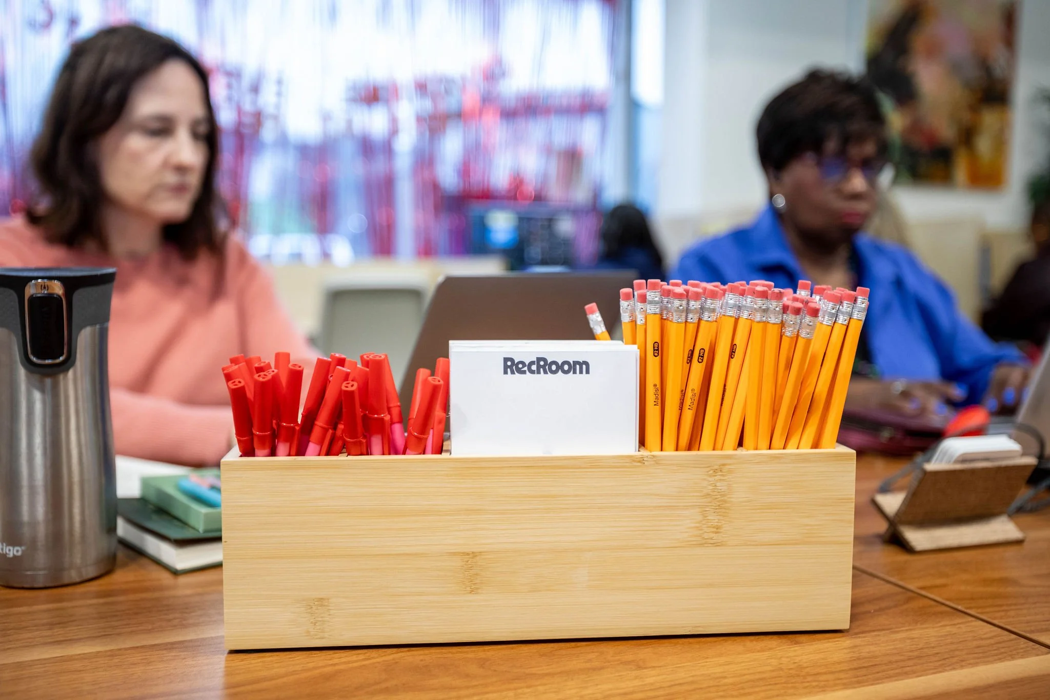 Desk organizer containing red markers, yellow pencils, and a white card that says 'RecRoom', with two women working in the background.