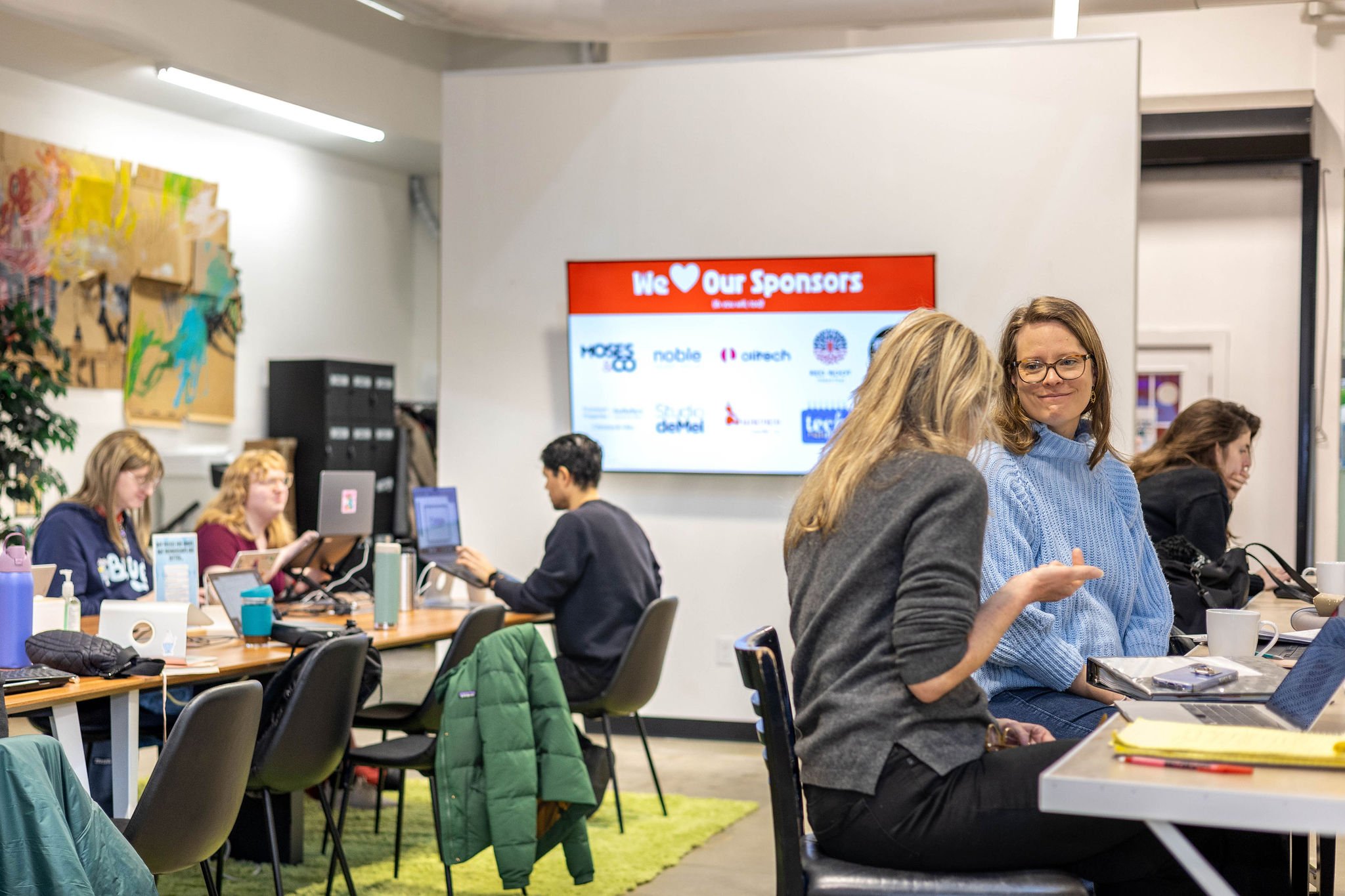 People in a meeting or collaborative space, some using laptops and tablets, with a large screen on the wall displaying sponsor logos and a red banner that reads 'We Heart Our Sponsors'.