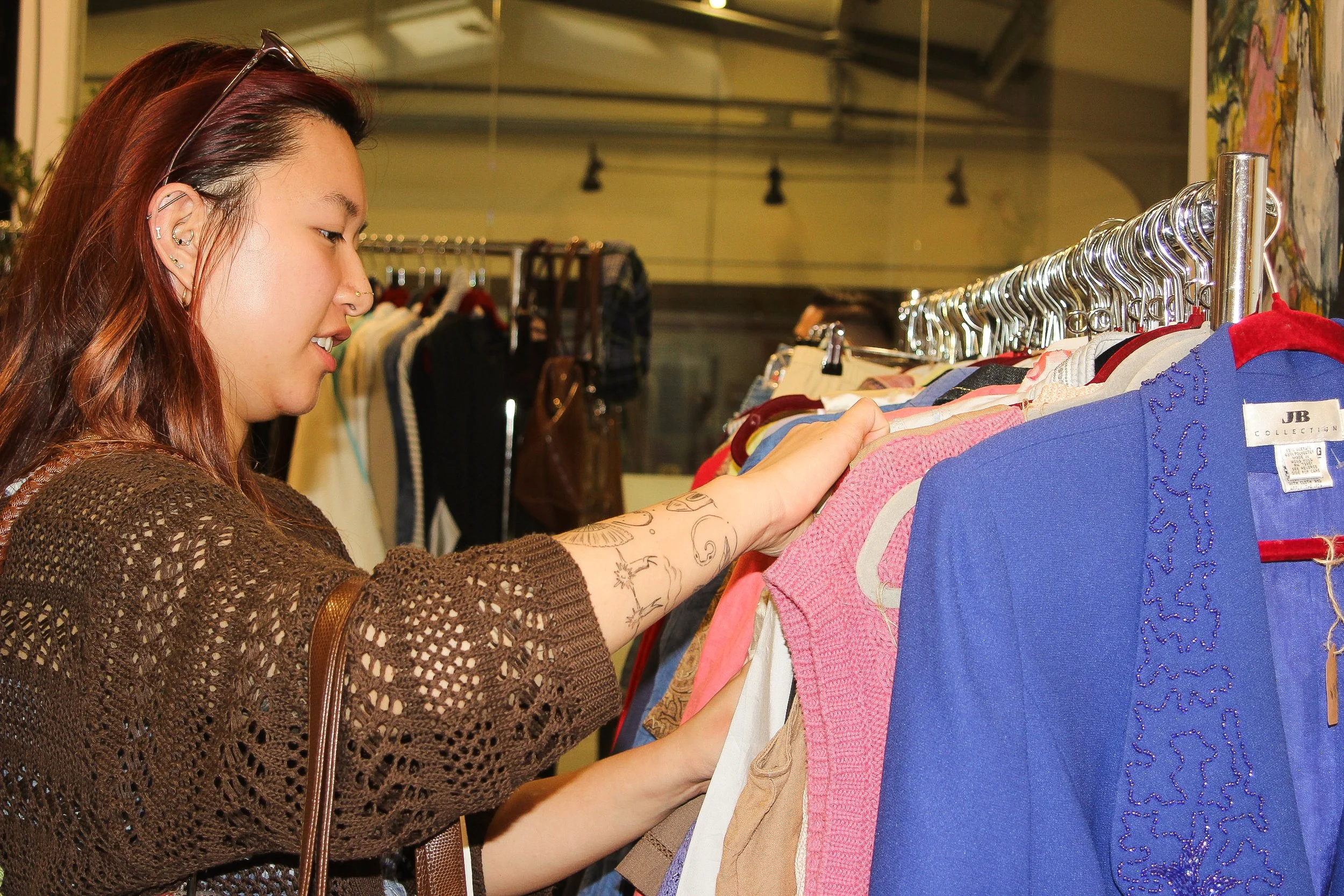 A young woman with tattoos on her arm is shopping for clothes at a thrift store, selecting an item from a rack of colorful clothing.