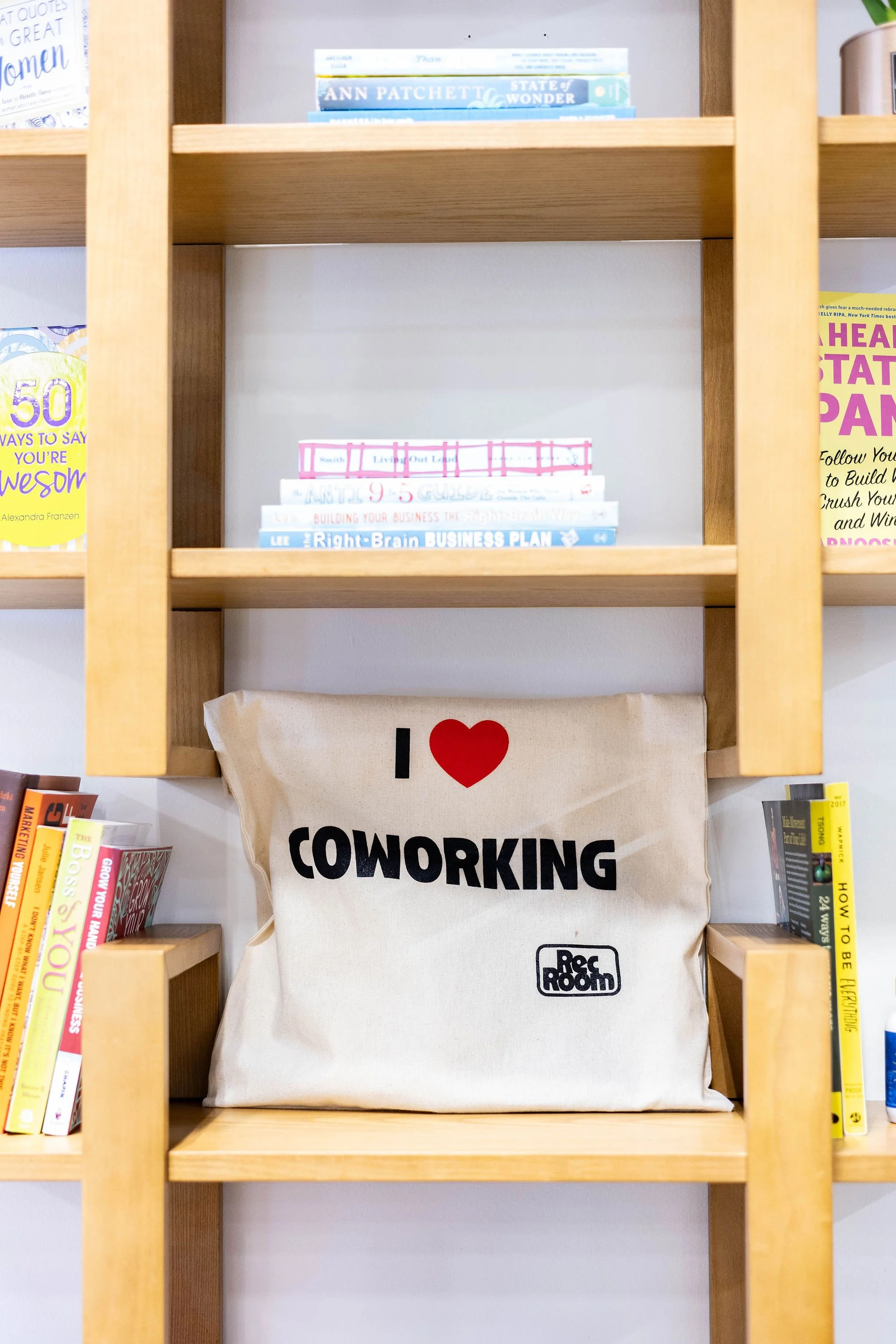 A beige tote bag with the text "I ❤️ COWORKING" and a small logo reading "Rec Room," placed on a wooden shelf, surrounded by books and displayed among other bookshelves.
