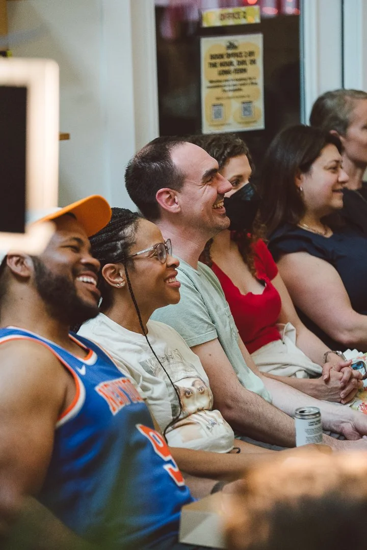 A group of people sitting in a row, smiling and enjoying a performance or event indoors. The group includes diverse individuals of different ages and ethnicities.
