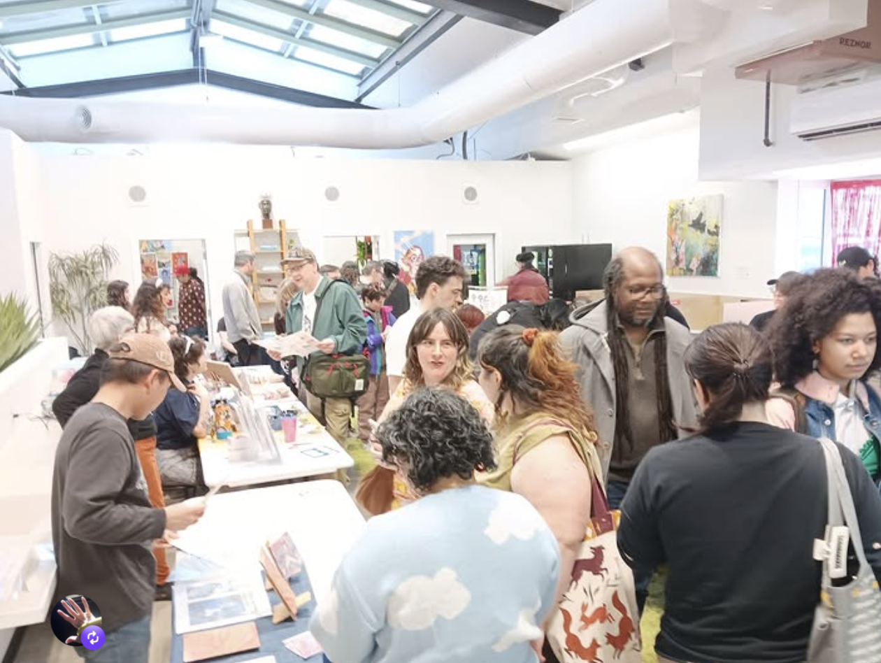 People gathered at tables in an art gallery or community space, viewing and discussing artwork displayed on the tables and walls, with a skylight above and various artwork in the background.