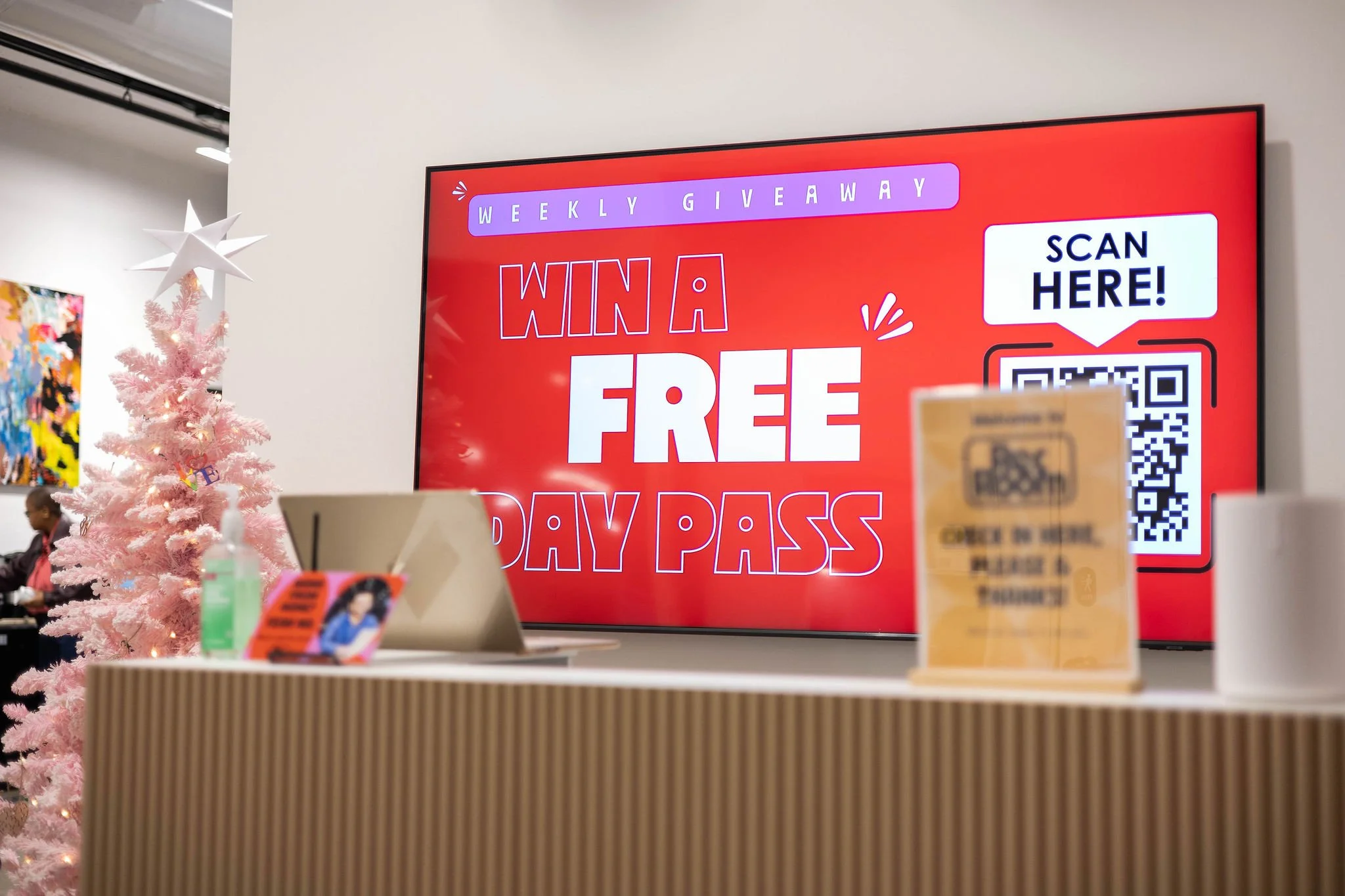 A digital screen displays a promotion for a weekly giveaway, offering a chance to win a free day pass. Next to the screen is a pink Christmas tree and a counter with a hand sanitizer bottle, a card, and a small sign.
