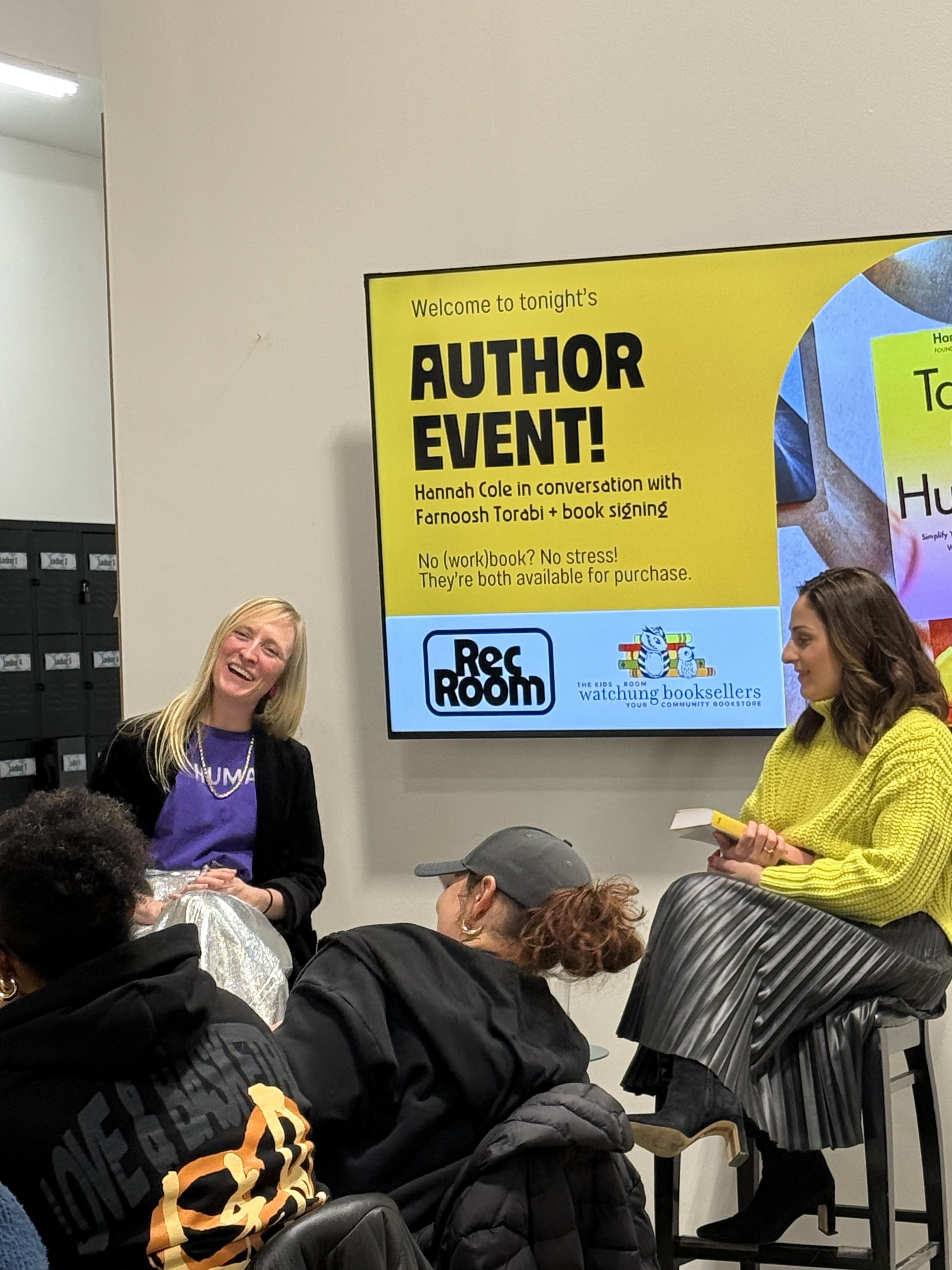 A group of people attending an author event with a large yellow sign in the background reading 'Welcome to tonight's Author Event! Hannah Cole in conversation with Fanoosh Torabi + book signing'.