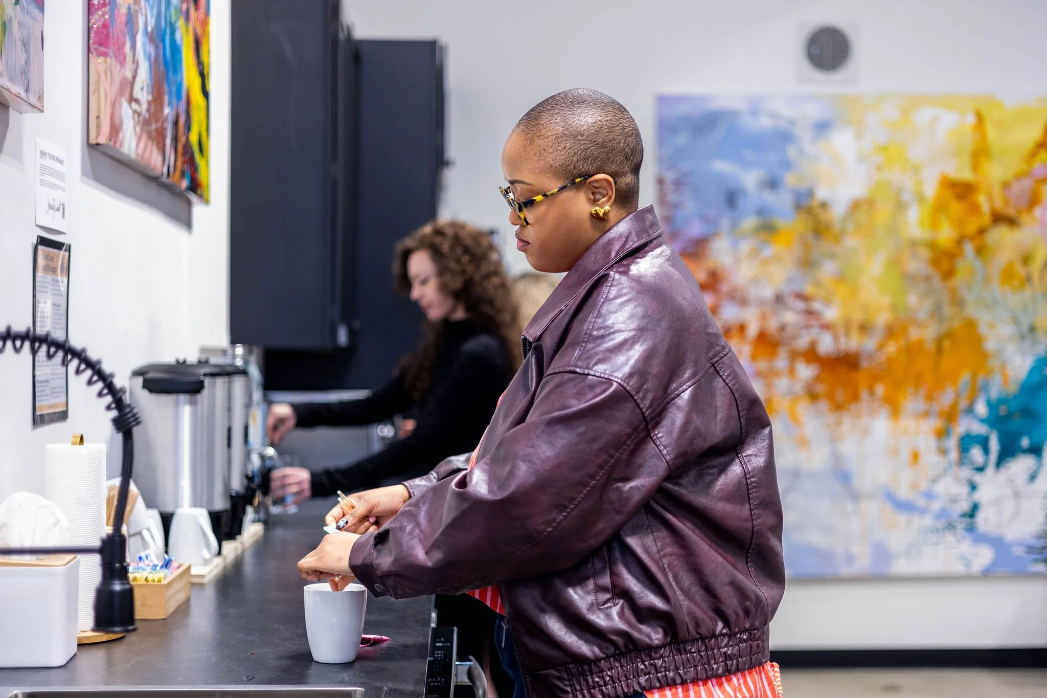 A woman with a shaved head wearing glasses, a purple leather jacket, and gold earrings, is standing at a counter in a cafe or office kitchen, pouring a beverage into a white mug. In the background, another woman with curly hair is preparing drinks, w