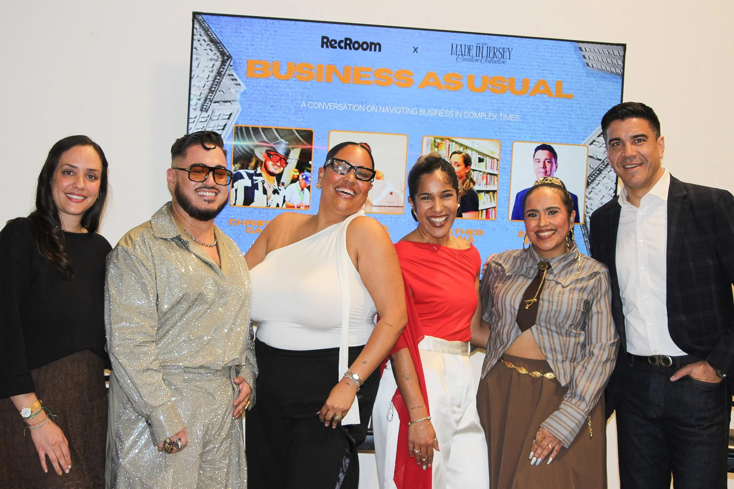Group of seven diverse people smiling and posing together at an indoor event, standing in front of a blue presentation screen with text about a business conversation.