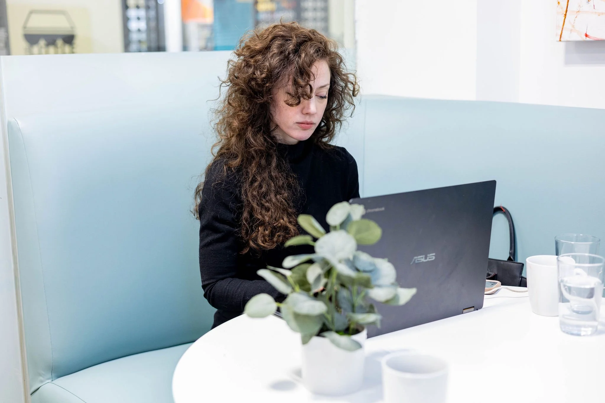 A woman with curly brown hair working on a black ASUS laptop at a white table, with a potted plant, cups, and glasses of water visible.