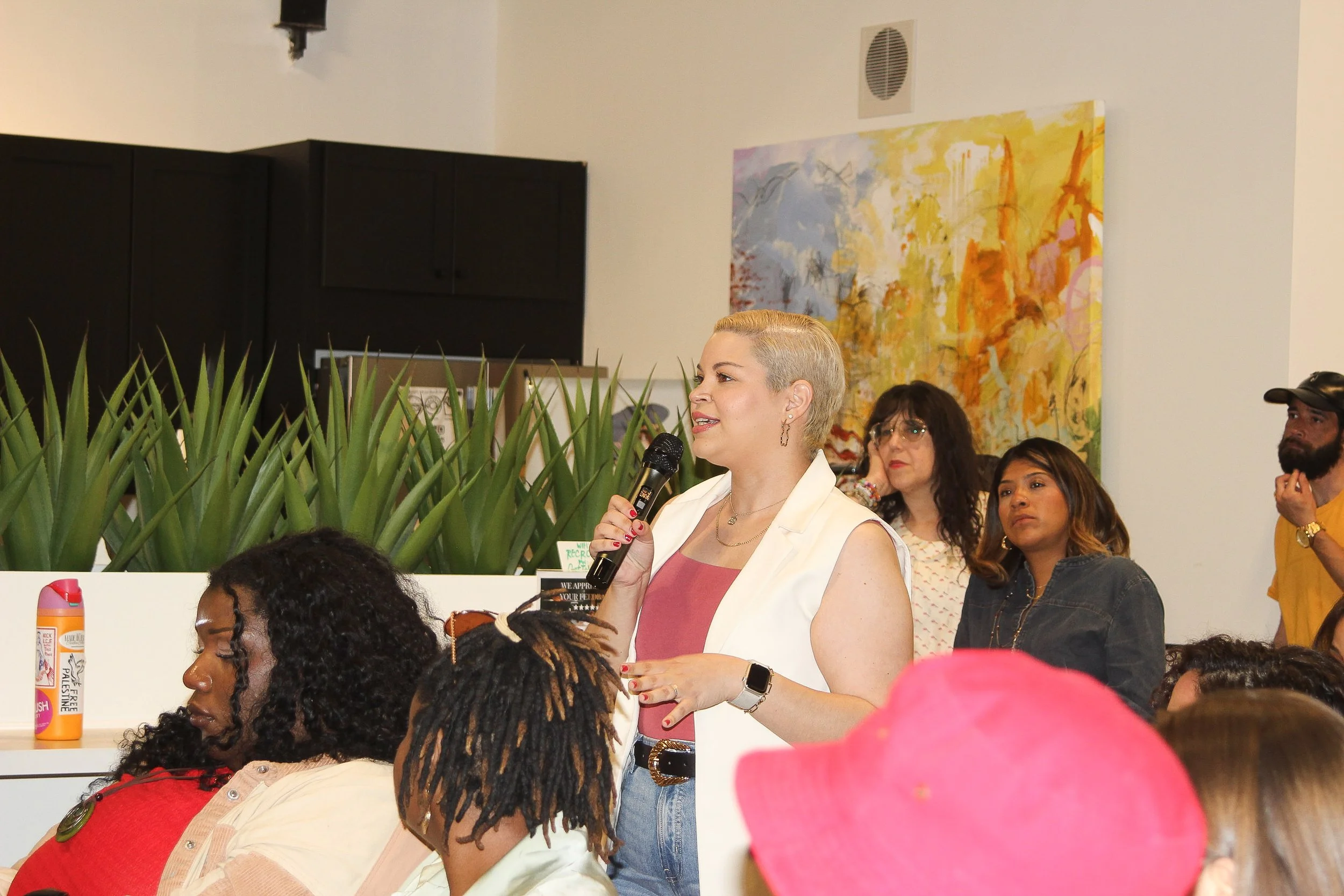 A woman with short blonde hair, wearing a pink top and white vest, holding a microphone, speaking to a crowd in an indoor setting decorated with abstract art and greenery.