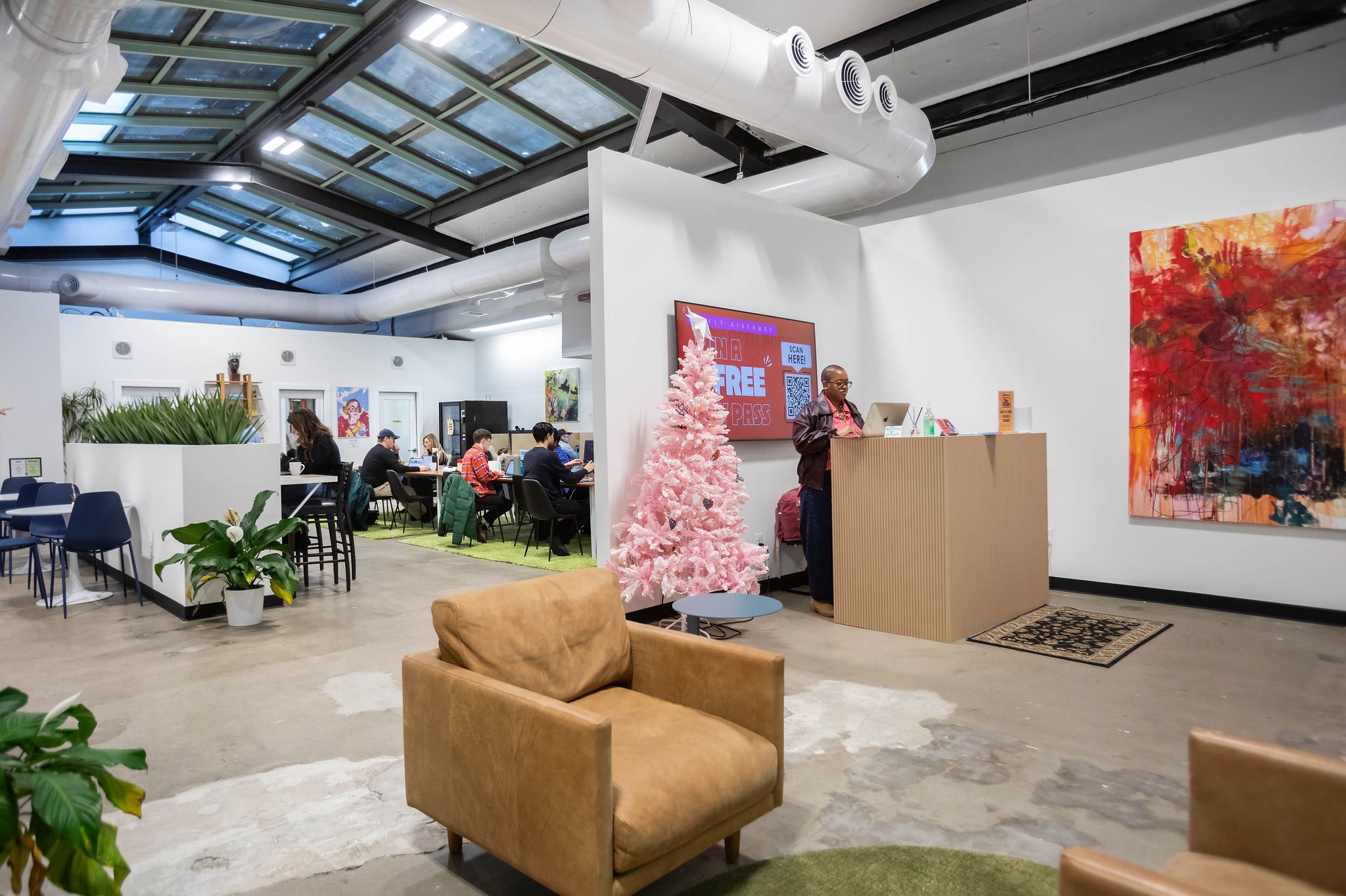 Interior of a modern coworking or community space with people working at tables. There is a pink Christmas tree near the reception desk with a man standing behind it, and a large colorful abstract painting on the wall. The space has a glass ceiling a