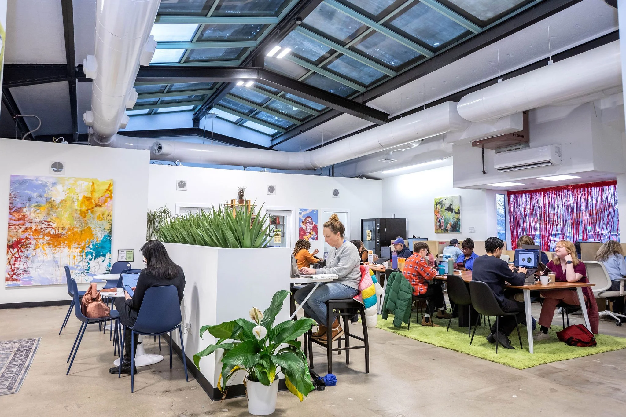 A modern co-working space with people working at desks and laptops, colorful abstract artwork on white walls, a large skylight ceiling, and plants, including a potted peace lily in the foreground.