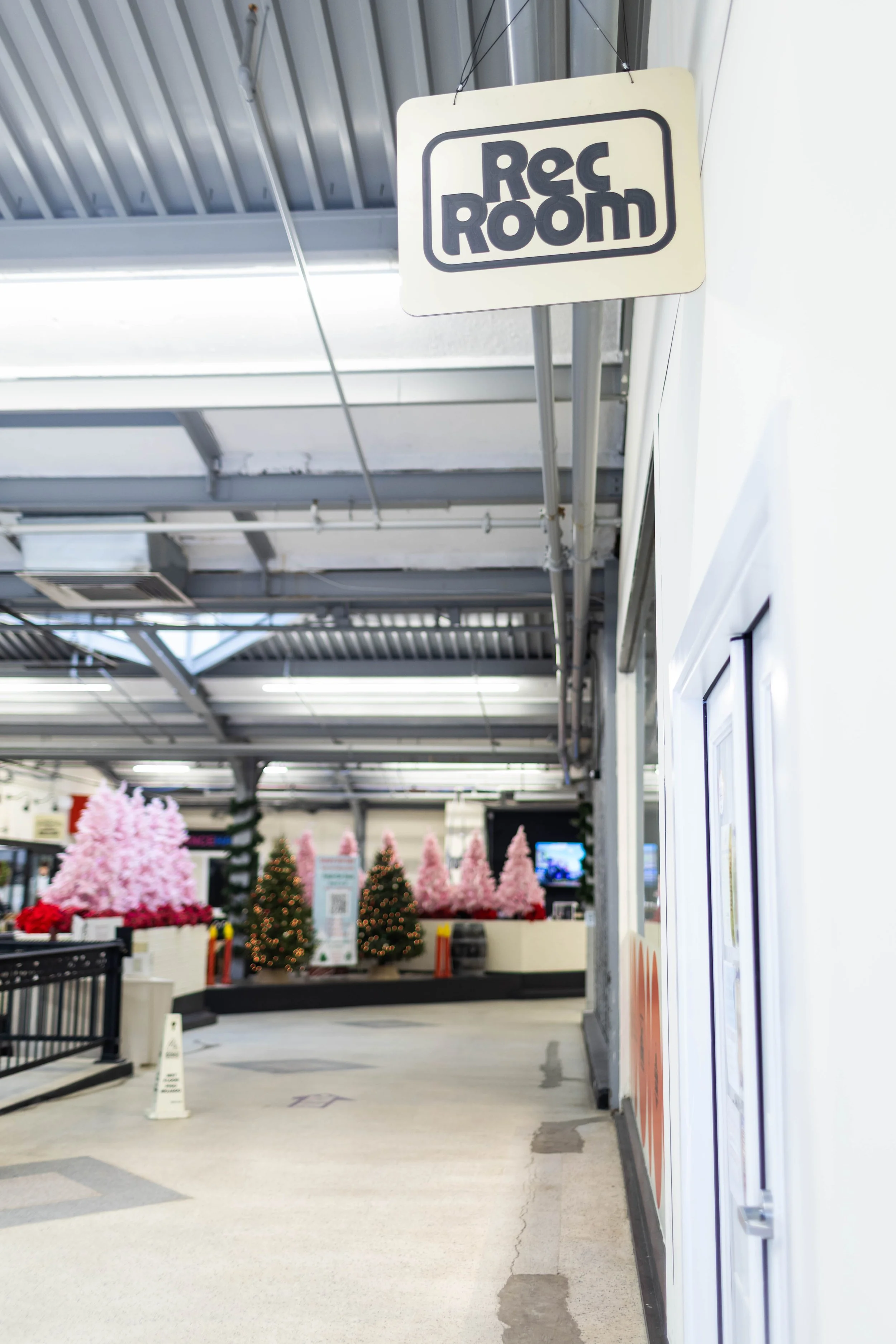 Photo of a sign that reads "Rec Room" hanging from the ceiling inside a shopping mall or store with Christmas trees and pink decorations in the background.