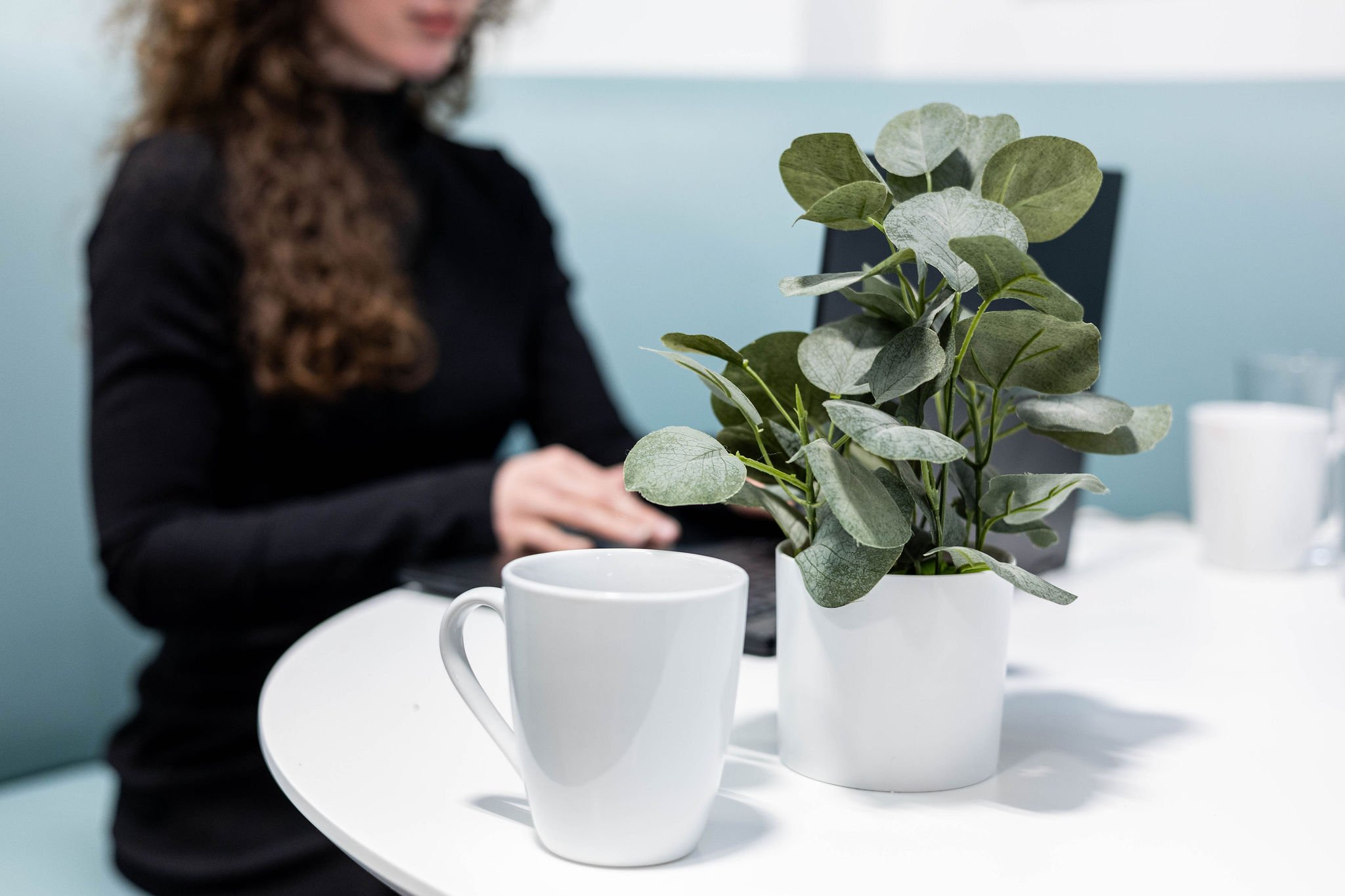 A woman with curly hair, dressed in black, sitting at a table with a laptop. On the table, there is a white mug and a potted plant with green leaves.