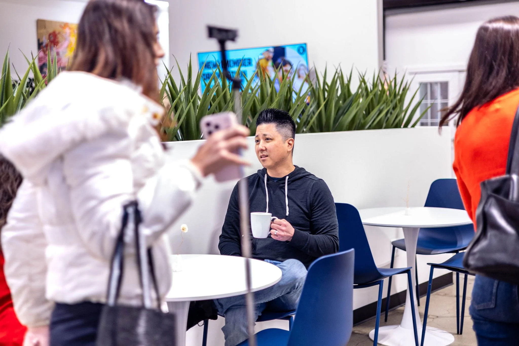 A man with short black hair, wearing a black hoodie, is seated at a white table in a modern cafe. He is holding a white mug and looking thoughtfully. Two women are in the foreground, blurred, one wearing a white jacket and holding a phone, the other 