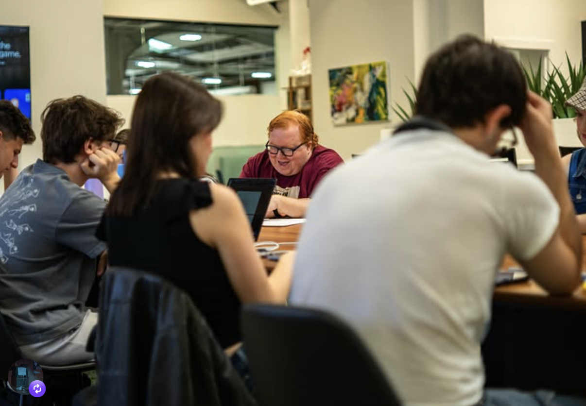 Group of young people sitting around a table, some working on laptops and others reading, in a room with colorful paintings and plants.