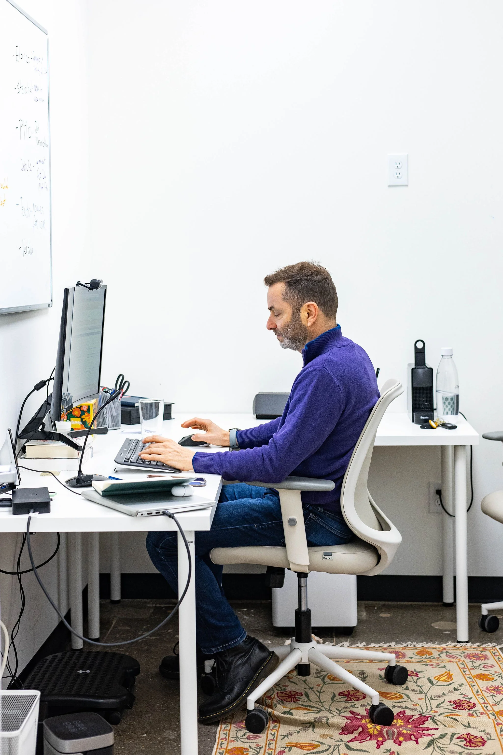 Man working at a white office desk with a computer, mouse, and various office supplies, sitting on an ergonomic chair in a minimalist office.