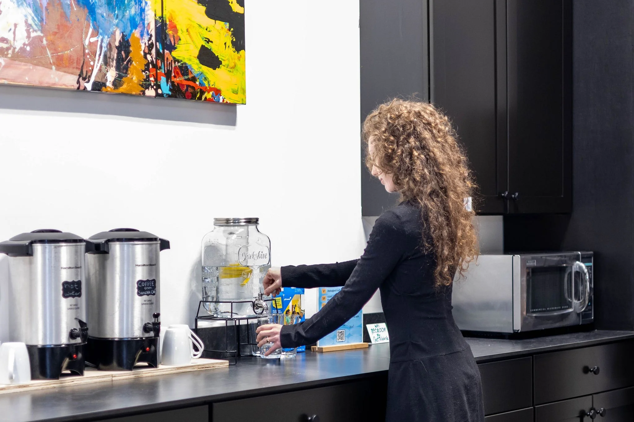 A woman with curly hair pouring water from a large glass jar into a glass on a black countertop in a break room with a colorful abstract painting on the wall.