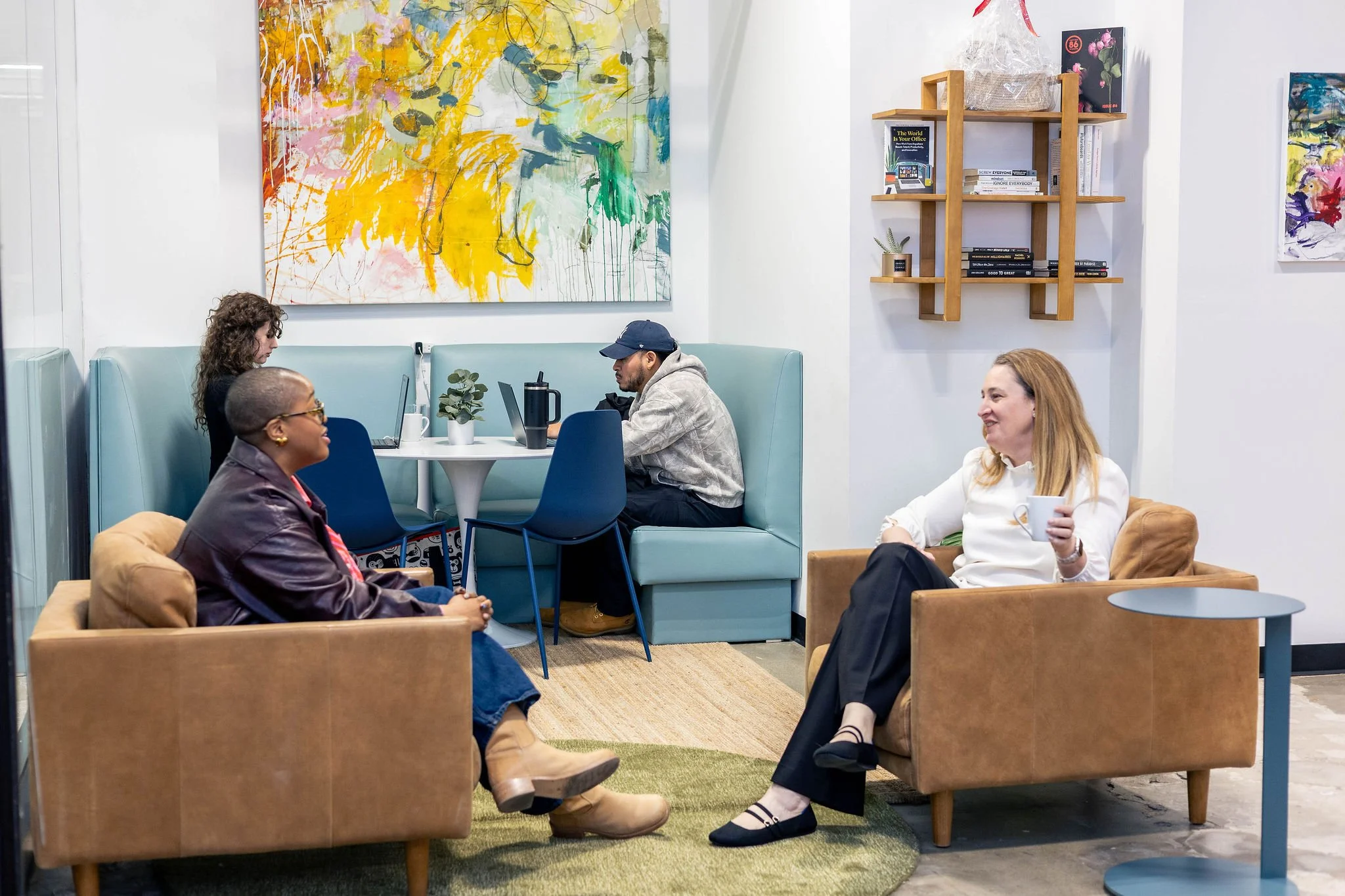 Four people sitting in a cozy lounge, engaged in conversation. Two women are seated in armchairs, one holding a coffee mug, and one man and woman sit at a small table with laptops and notebooks. The room has colorful abstract artwork on the walls and