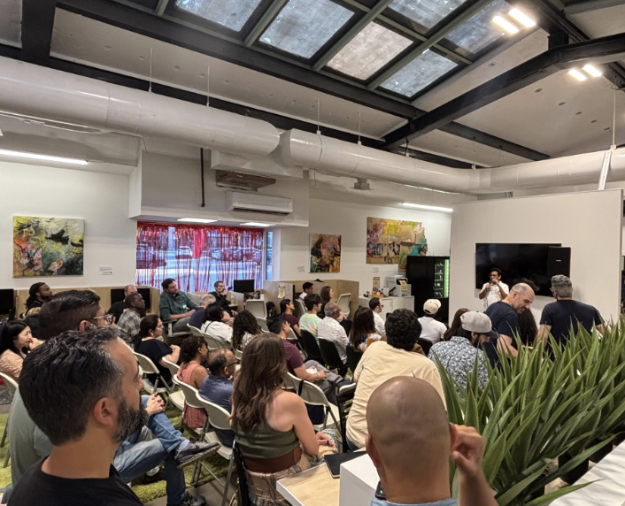 An indoor event with a large audience seated and watching a speaker at the front of the room. The room has a high ceiling with skylights, artwork on the walls, and a large flat-screen TV. Some attendees are taking photos or listening attentively.