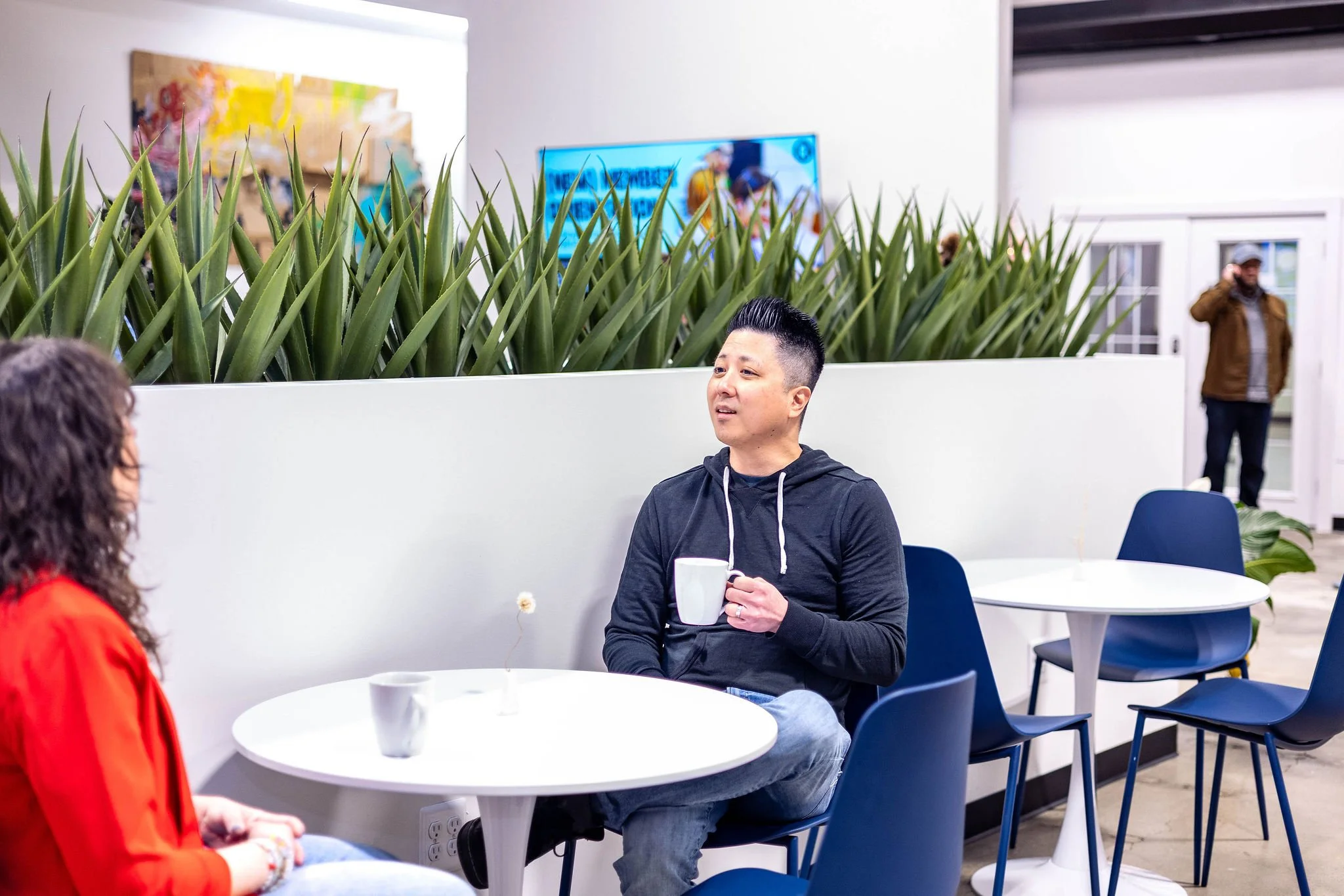 Two people sitting at a white table in a modern cafe, engaged in conversation. One person is a woman with curly hair wearing a red jacket, and the other is a man with short black hair, wearing a black hoodie, holding a mug. In the background, a man i