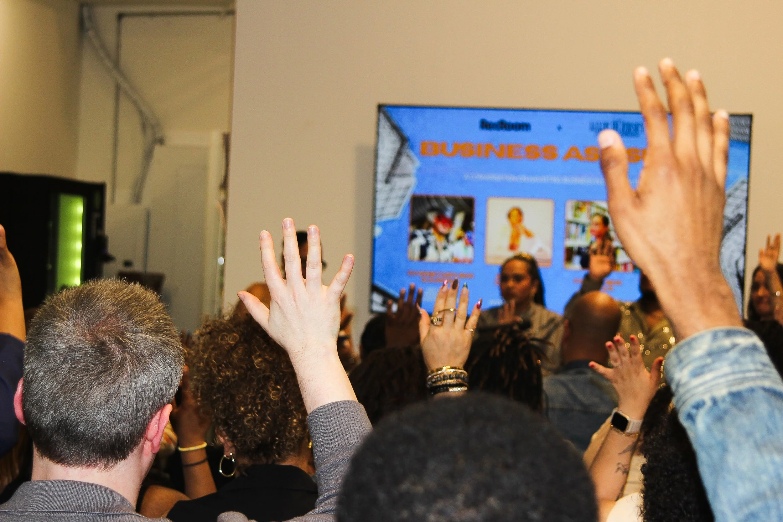 Audience raising hands during conference or presentation in a room with a digital screen displaying a presentation titled 'Business As...'.