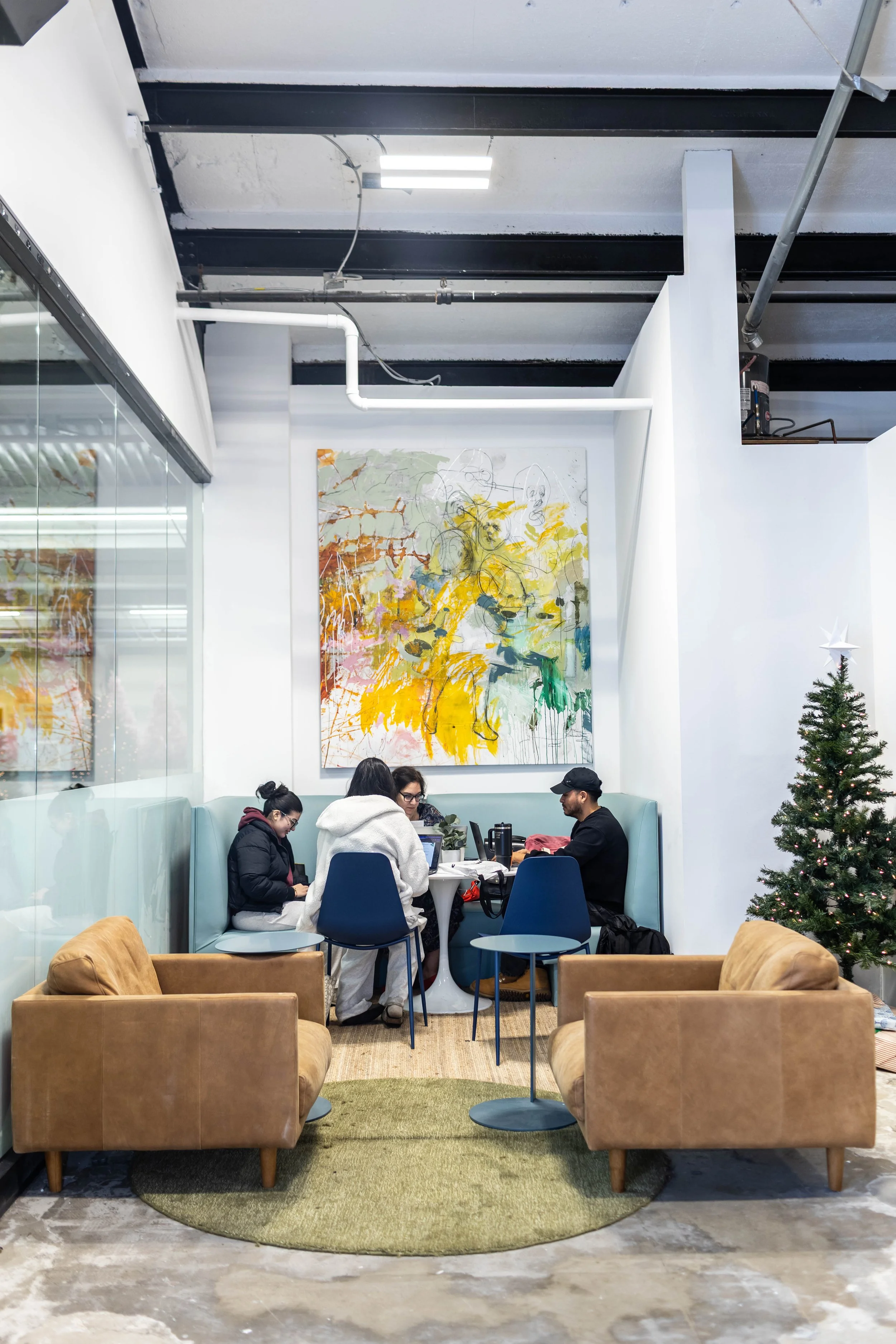 Interior of a modern cafe or lounge with four people sitting at a table, a large colorful abstract painting on the wall, a decorated Christmas tree to the right, and a mix of leather and fabric seating.