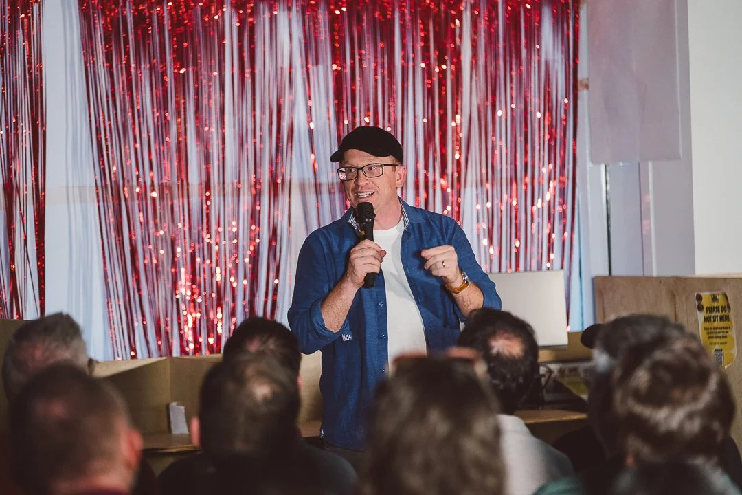 A man in glasses, a black cap, and a blue jacket speaking into a microphone in front of a decorated red and white curtain at a comedy or public event, with an audience in the foreground.