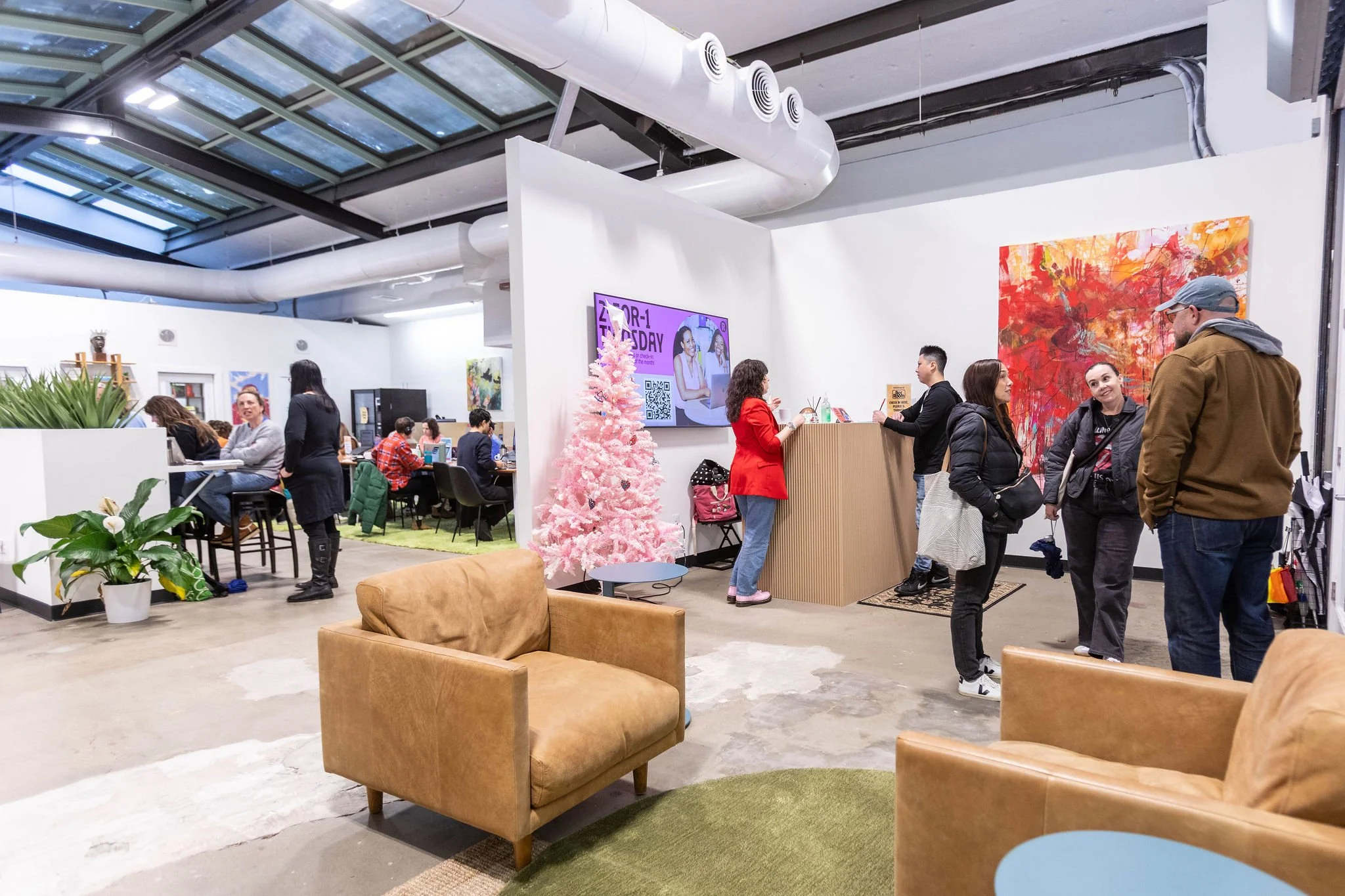 People standing in line at a reception desk in a modern, open-concept office or coworking space with artwork, pink Christmas tree, and casual seating.