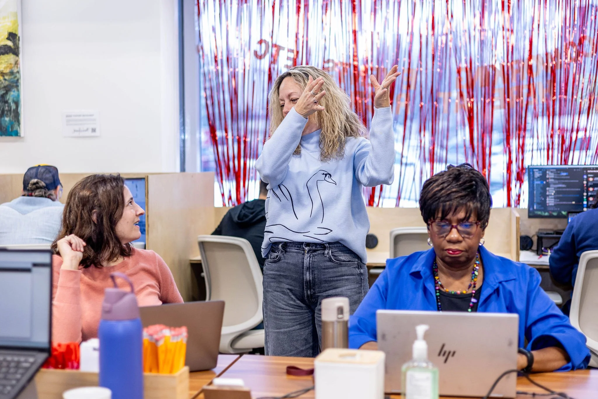 A woman with curly blond hair standing and gesturing with her hands while talking in a busy office. Two women are seated at desks, one with brown hair and the other with short black hair, working on laptops.