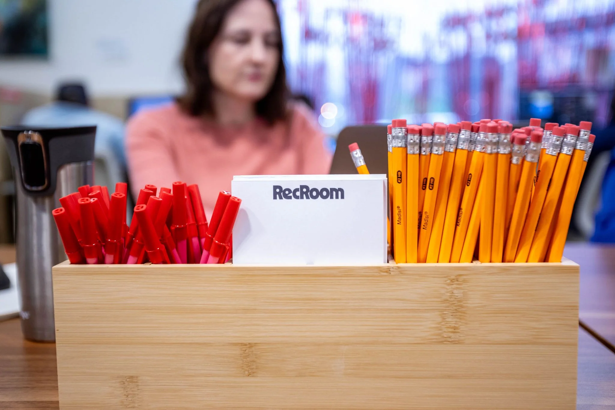 A wooden organizer on a desk holding red pens, yellow pencils with erasers, and a white sign with black text that says 'RecRoom.' In the background, a woman with shoulder-length brown hair wearing a pink top is working on a laptop in a busy office en