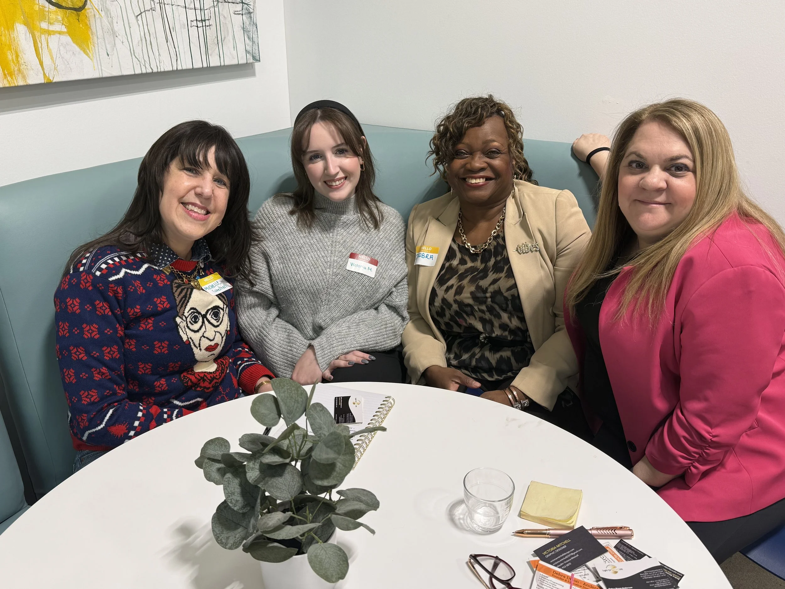 Four women sitting closely together at a round white table in a room with cream-colored walls. They are smiling and looking at the camera. The table has a small plant, a glass of water, a yellow notepad, a pair of glasses, and some business cards. Th