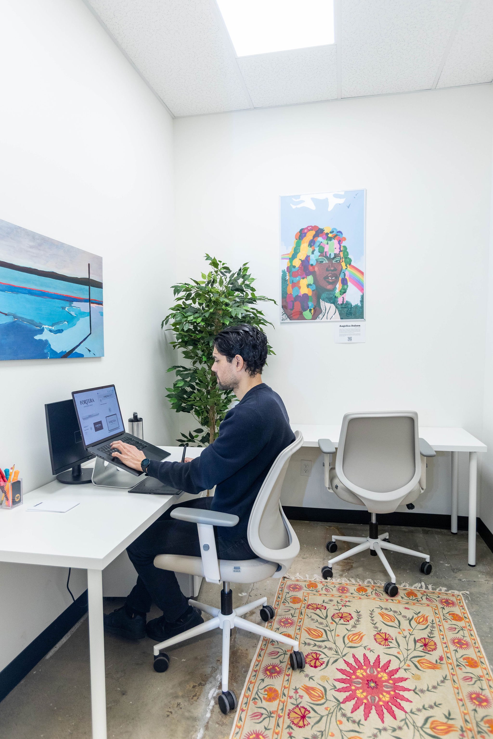 A man working at a desk in an office with two paintings on the white wall, a potted plant, an empty chair, and a colorful rug on the concrete floor.