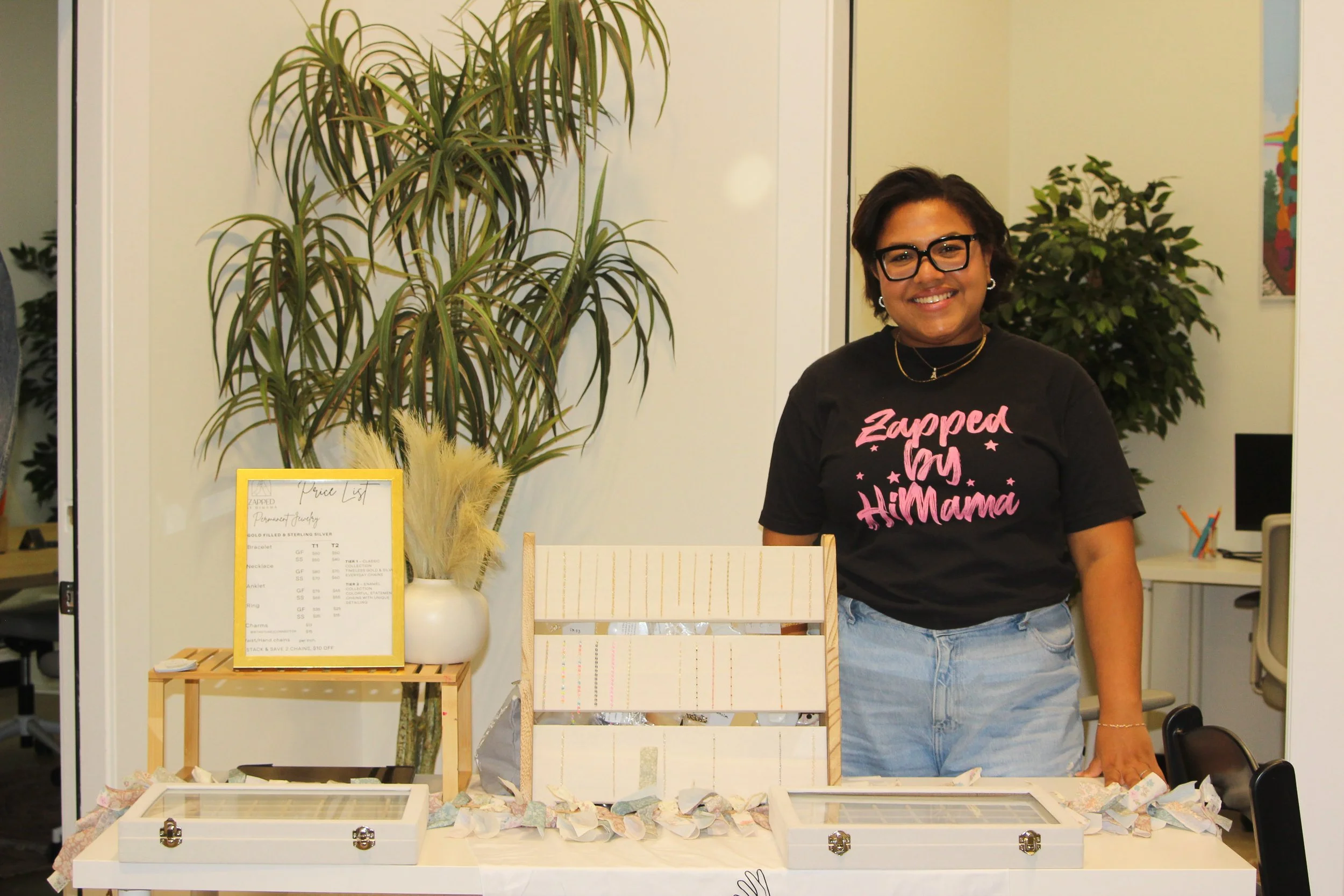 Smiling woman with glasses standing behind a table displaying jewelry and gemstones, with large potted plants and a price list in the background.
