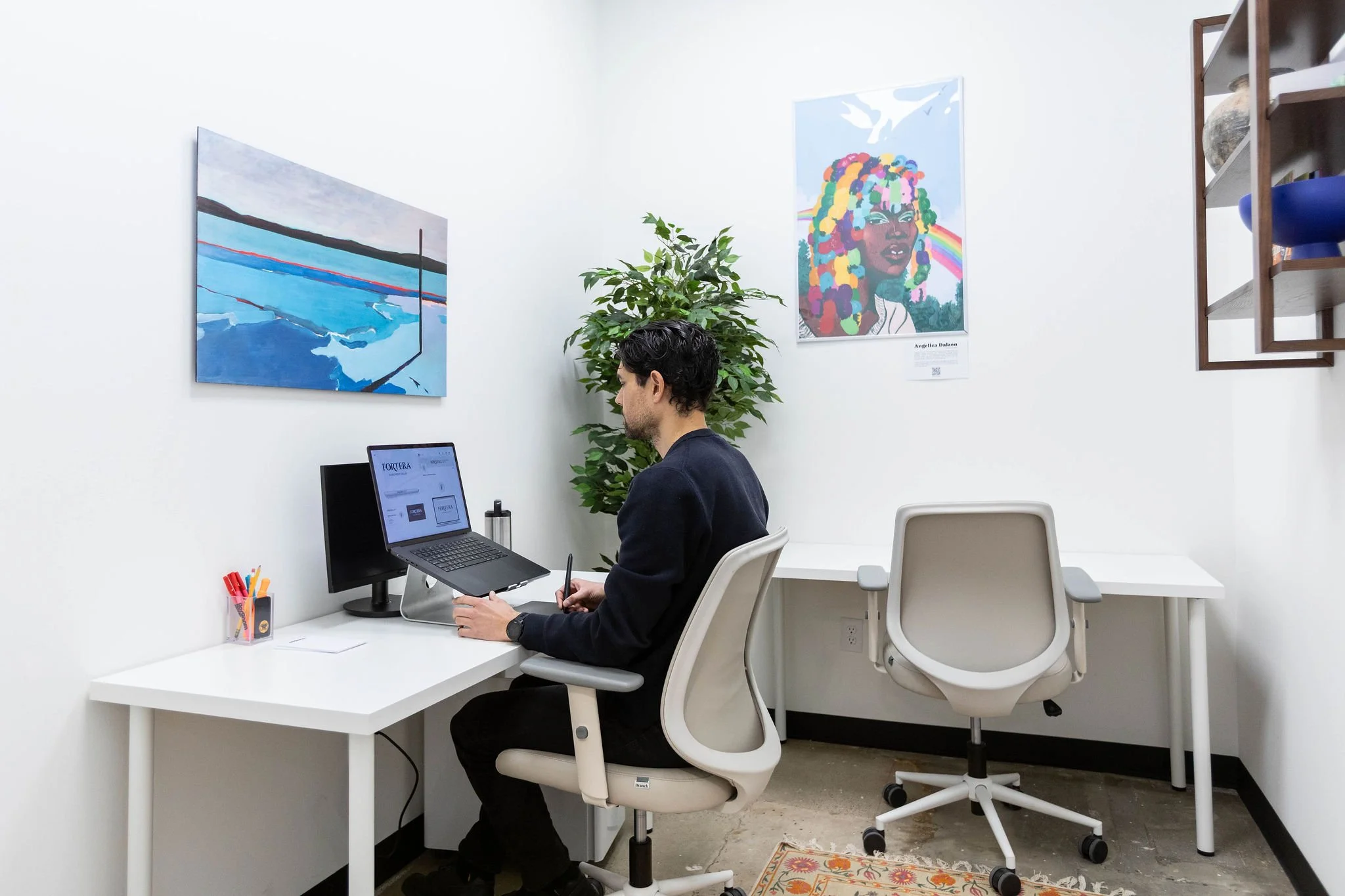 A man sitting at a white desk with a laptop in a small, minimalist office with white walls. Artworks hang on the wall behind him, including a colorful portrait and an abstract landscape. There is a large green plant and shelves with decorative items.