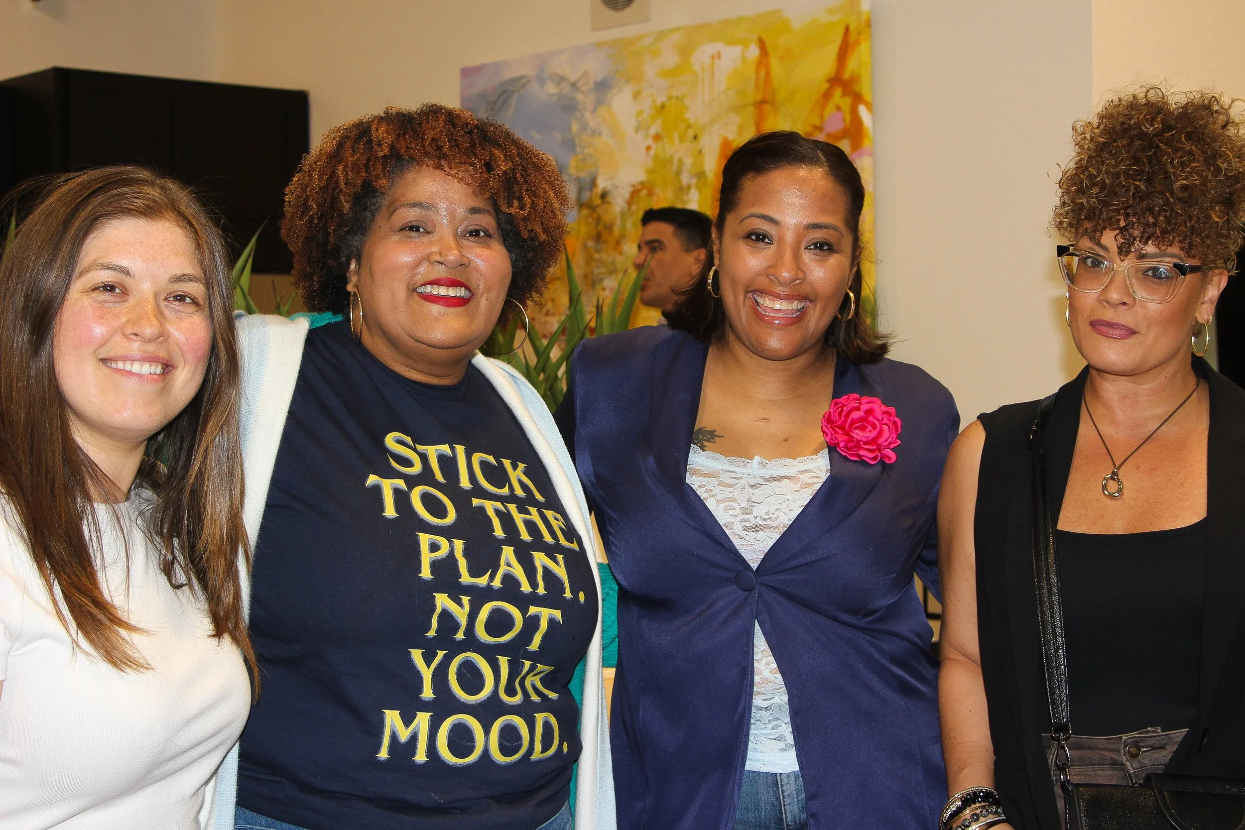 Group of four women smiling at a social gathering, standing in front of a colorful abstract painting and a beige wall.
