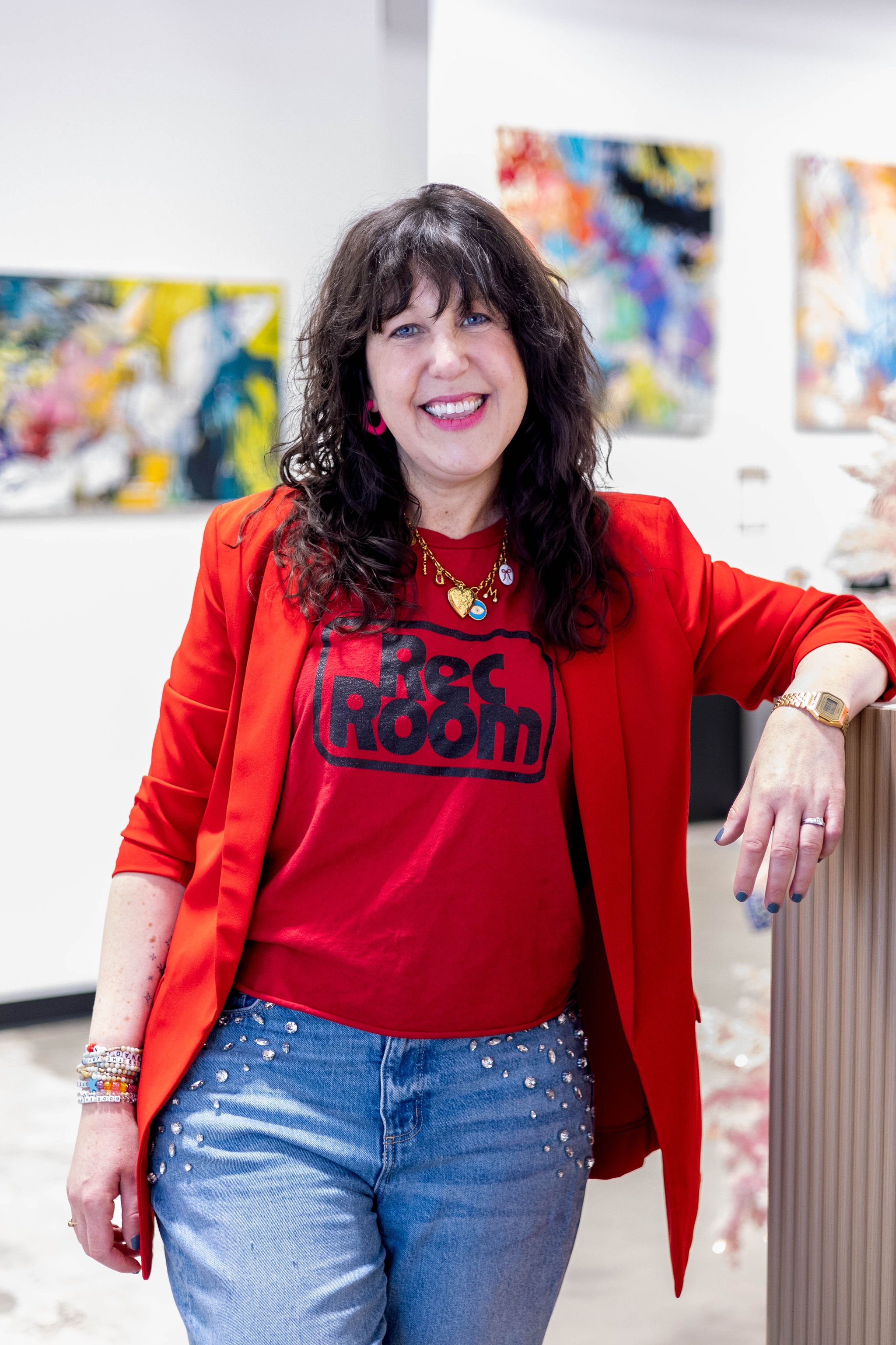 A woman with wavy dark hair wearing a red blazer, red t-shirt with black text, and light blue jeans decorated with rhinestones, standing in front of colorful abstract paintings in an art gallery.