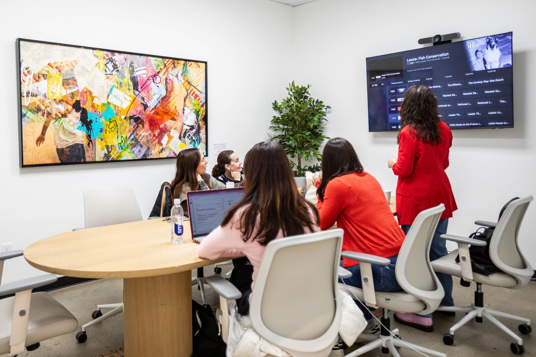 A group of women in a meeting room, one woman standing and presenting to others seated around a round table, with a large colorful art piece on the wall and a screen displaying a TV guide.